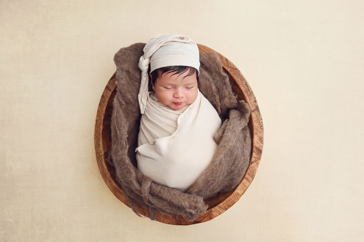 Newborn baby peacefully sleeping in cream swaddle blanket inside brown wooden bowl on beige backdrop. Fresno Newborn Photographer, Christina