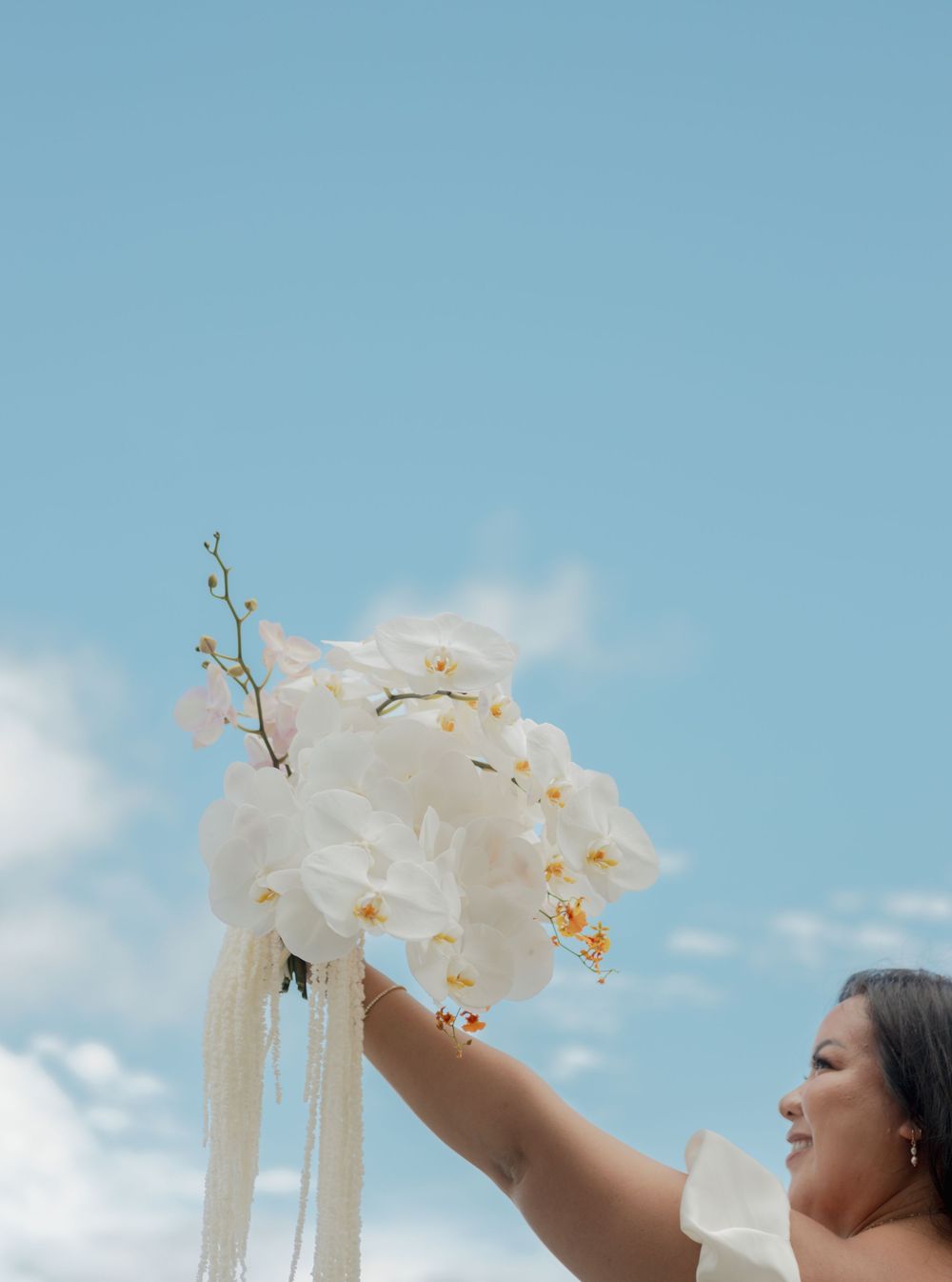 A hand raises a bouquet of white orchids against a bright blue sky with wispy clouds.