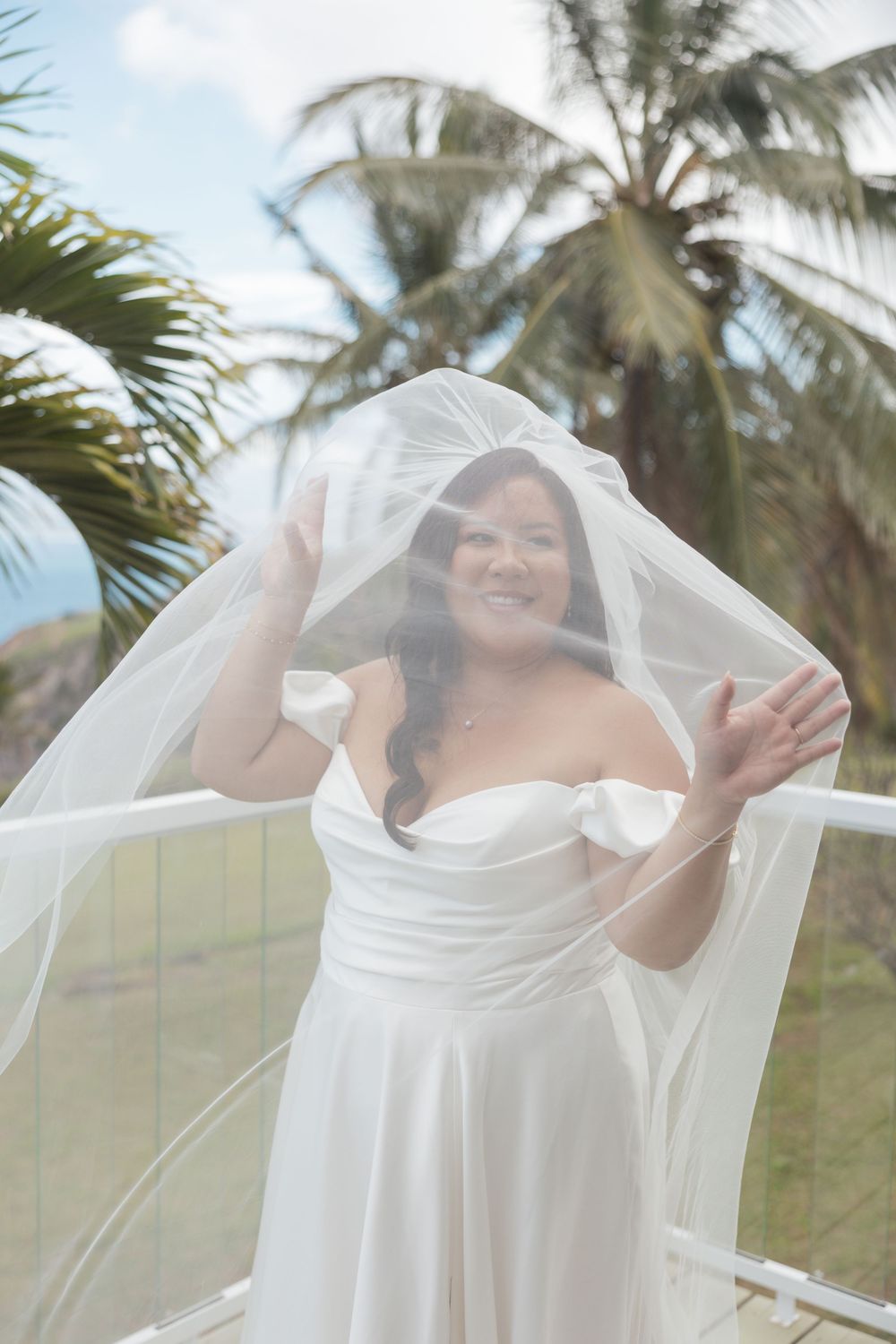 A bride in a flowing white off-shoulder wedding dress playfully lifts her veil against a tropical palm tree backdrop.