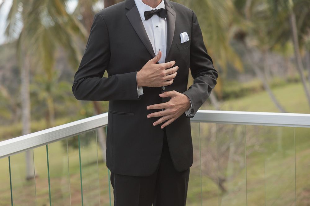 Close-up view of a black formal tuxedo with bow tie shown on a balcony with palm trees in background.