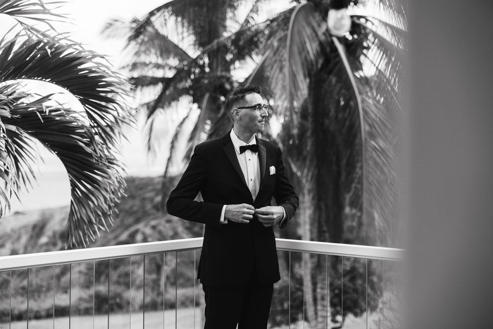 A groom in a classic black tuxedo stands on a tropical balcony with palm trees in the background in black and white.