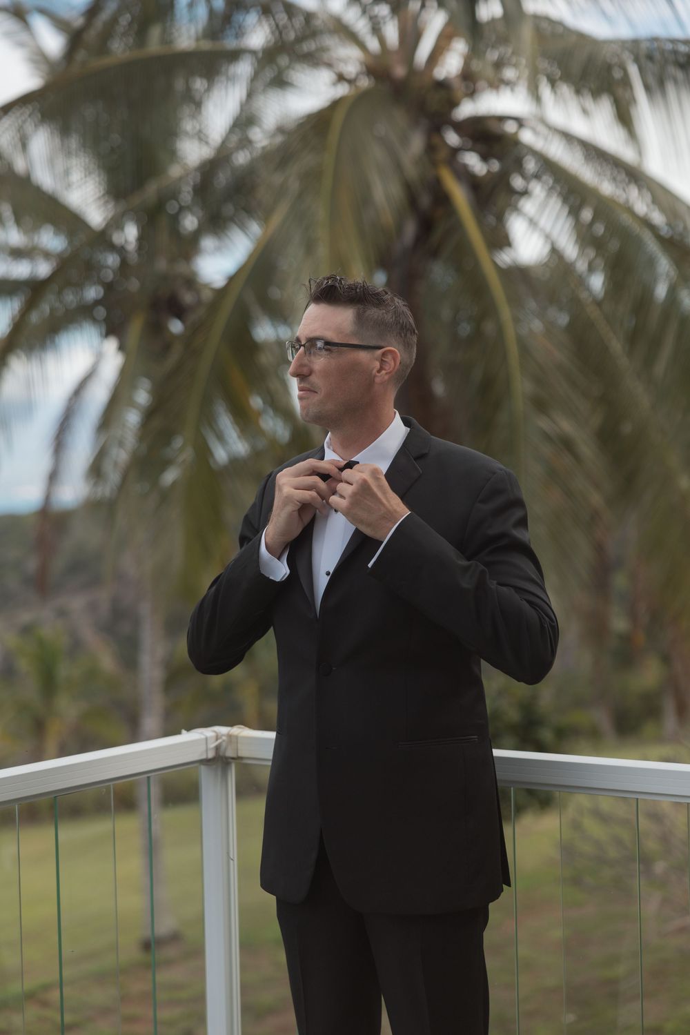 A well-dressed person in a black suit adjusts their bowtie while standing on a balcony with palm trees in the background.