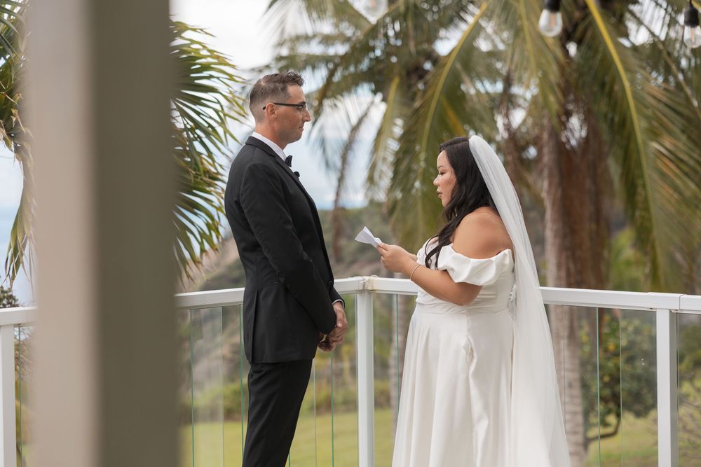 A couple exchanges rings during a tropical outdoor wedding ceremony on a white balcony with palm trees in the background.