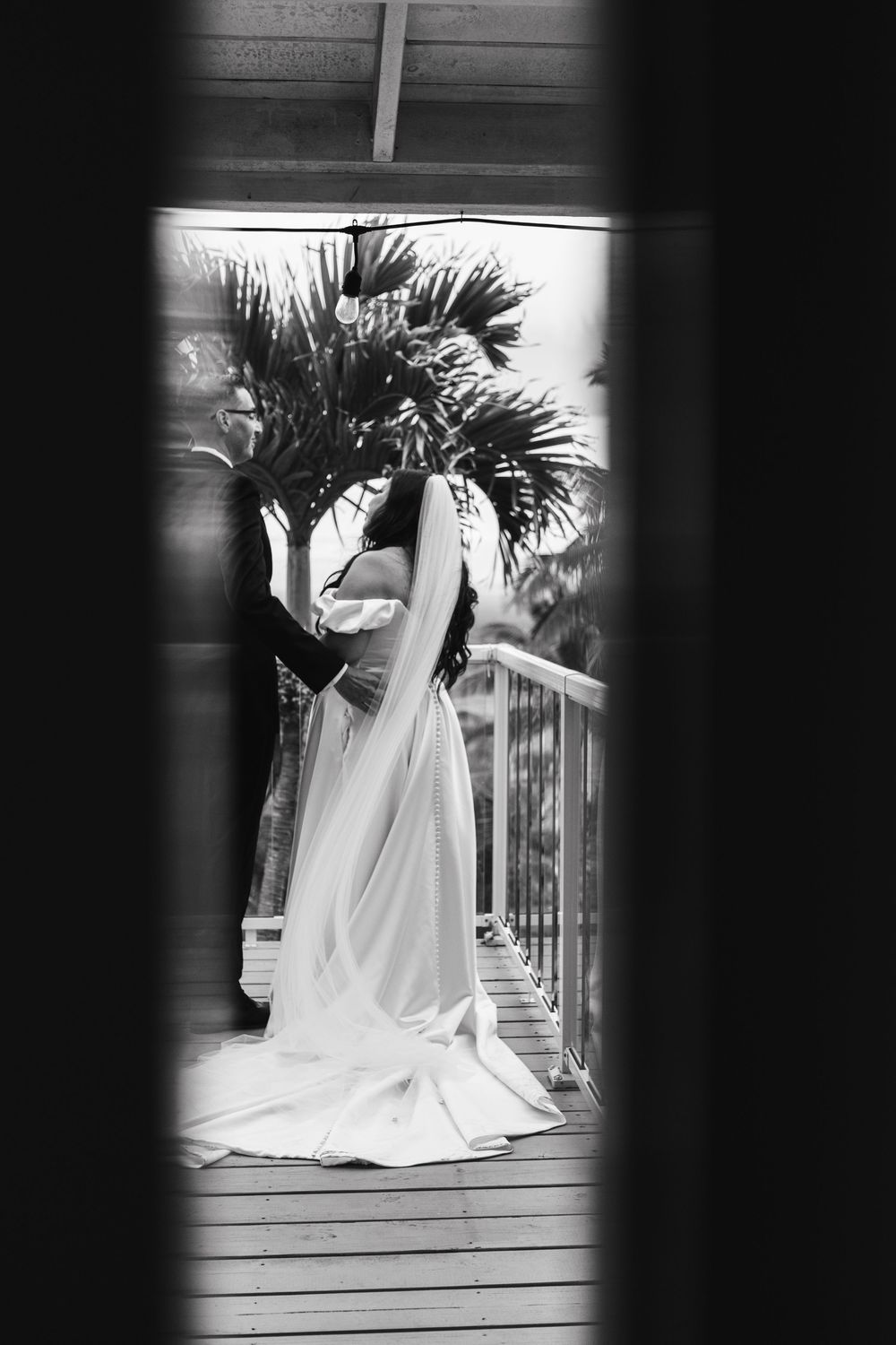 A black and white artistic photo of a bride in silhouette standing on a deck with palm trees.