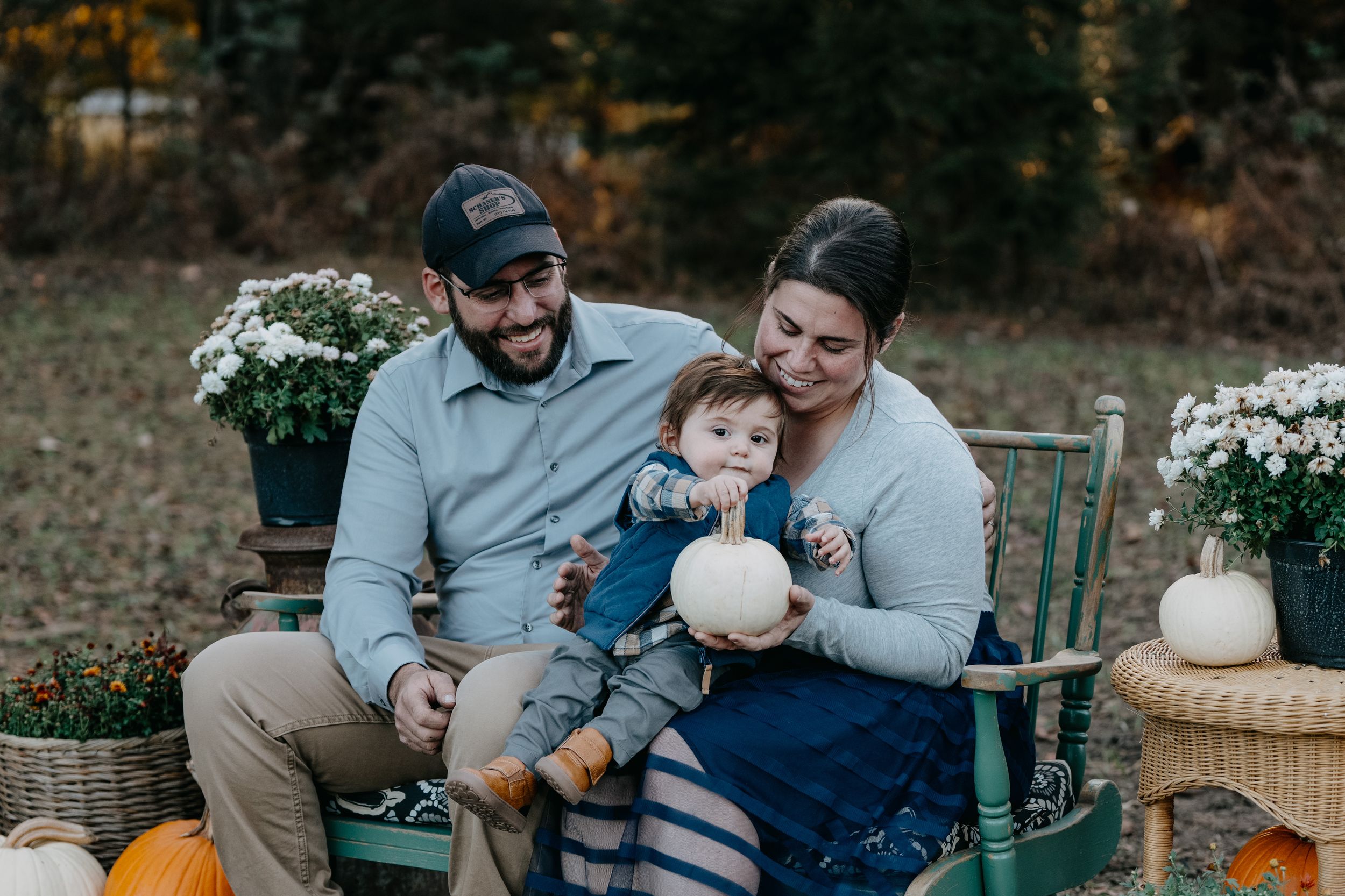 A family sits together on a rustic wooden bench surrounded by autumn pumpkins and white floral arrangements in the garden.