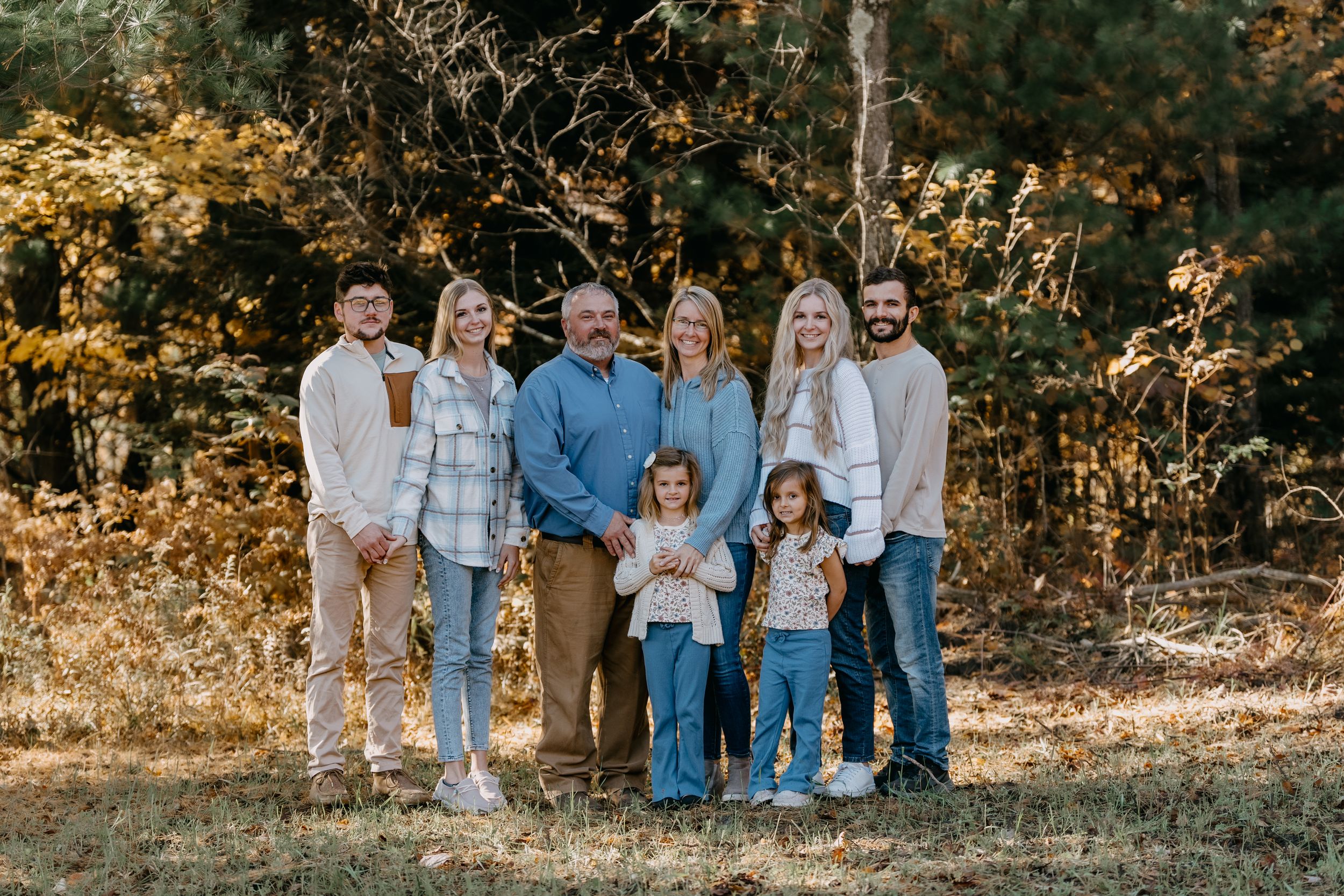 A group stands together outdoors in casual clothing against a natural autumn backdrop with golden foliage.