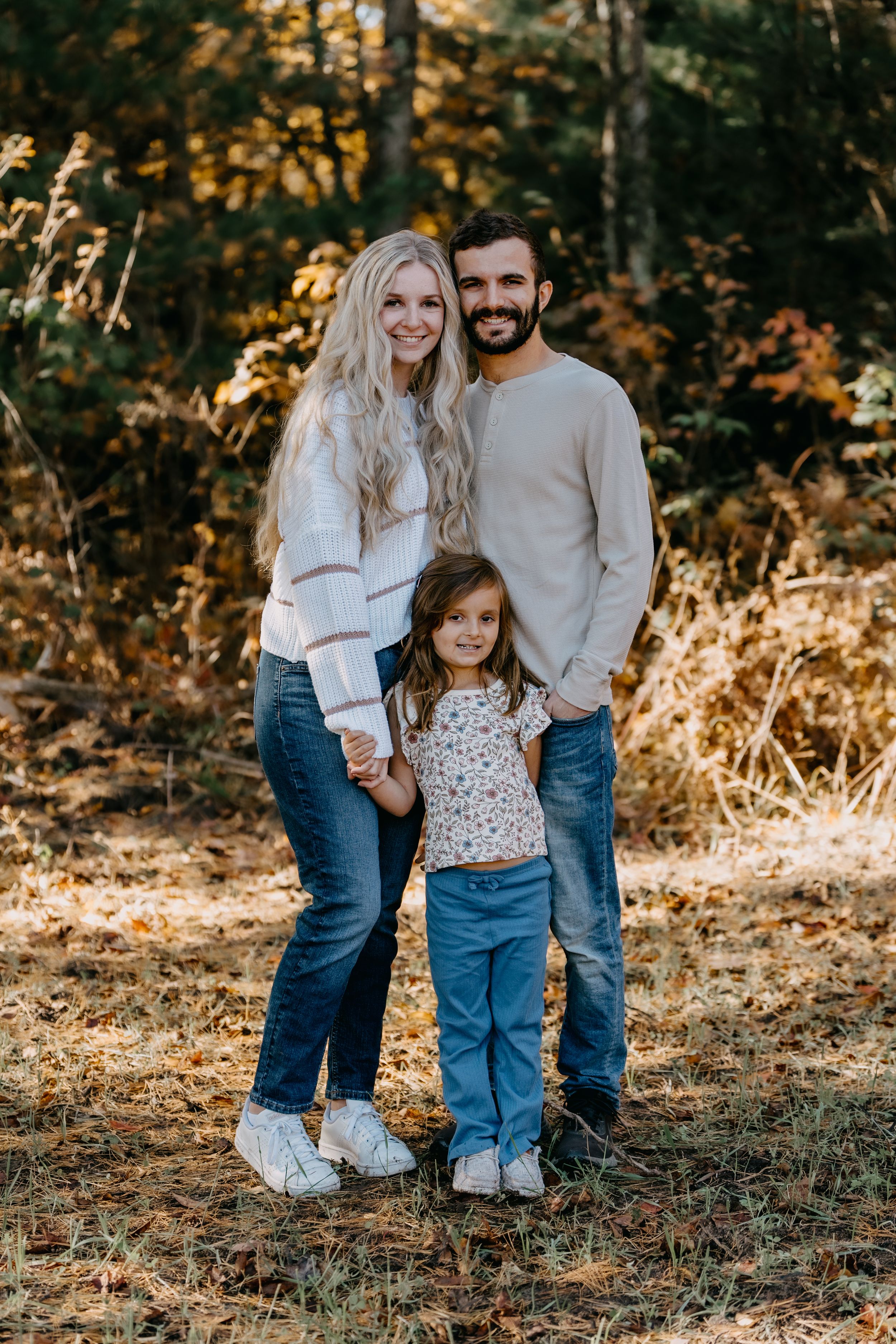 Family in matching cream sweaters poses together in autumn forest with golden sunlight filtering through trees.