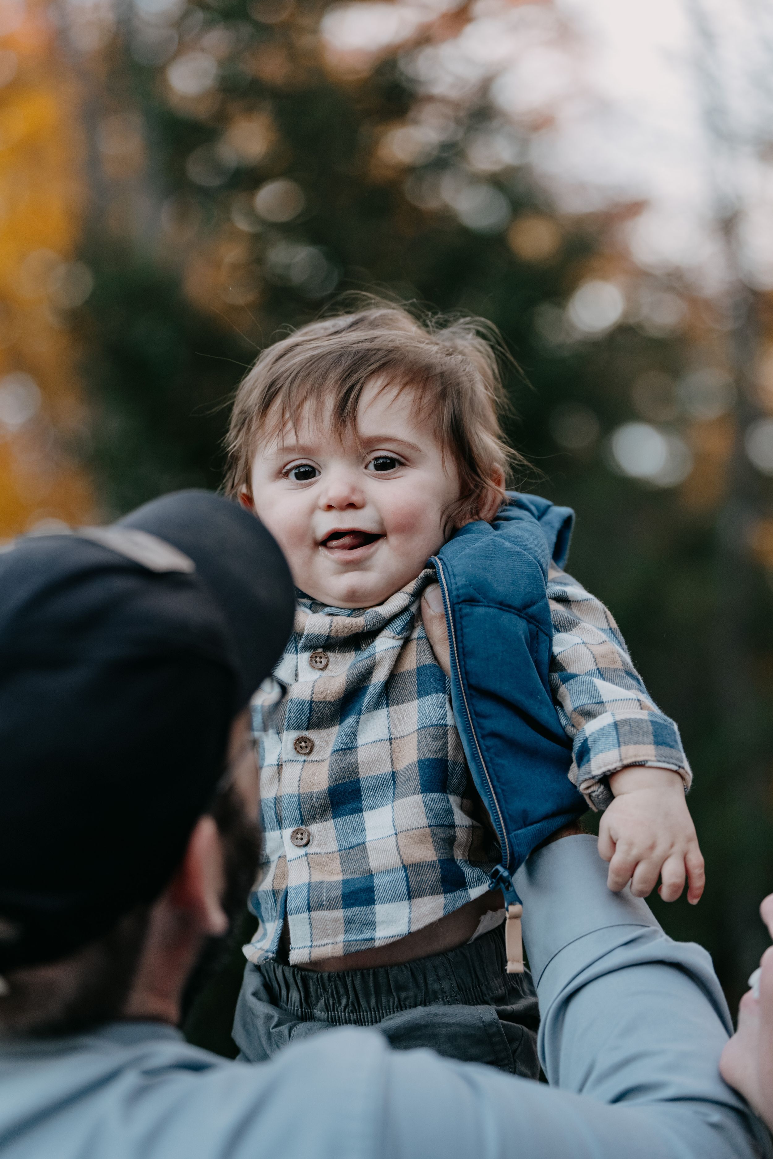 A young child wearing a blue and white plaid shirt and vest smiles joyfully while being lifted up outdoors.