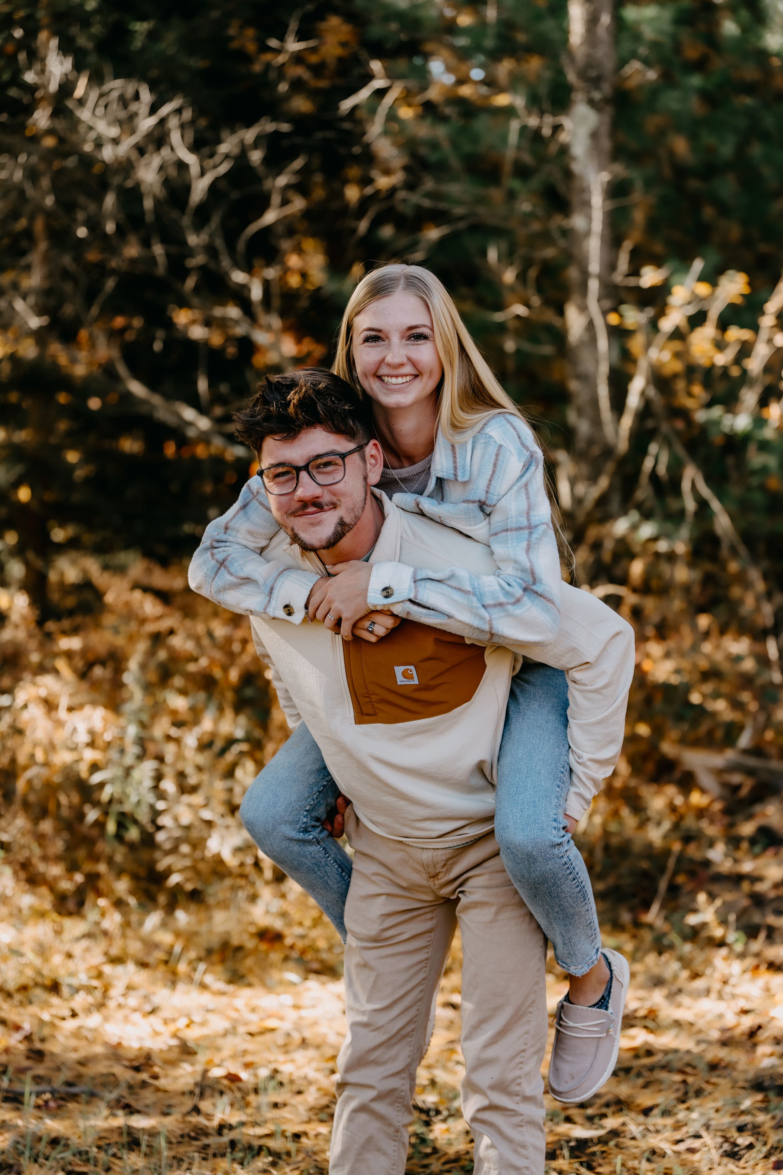 Playful couple enjoying a piggyback ride together in a sun-dappled autumn forest setting.