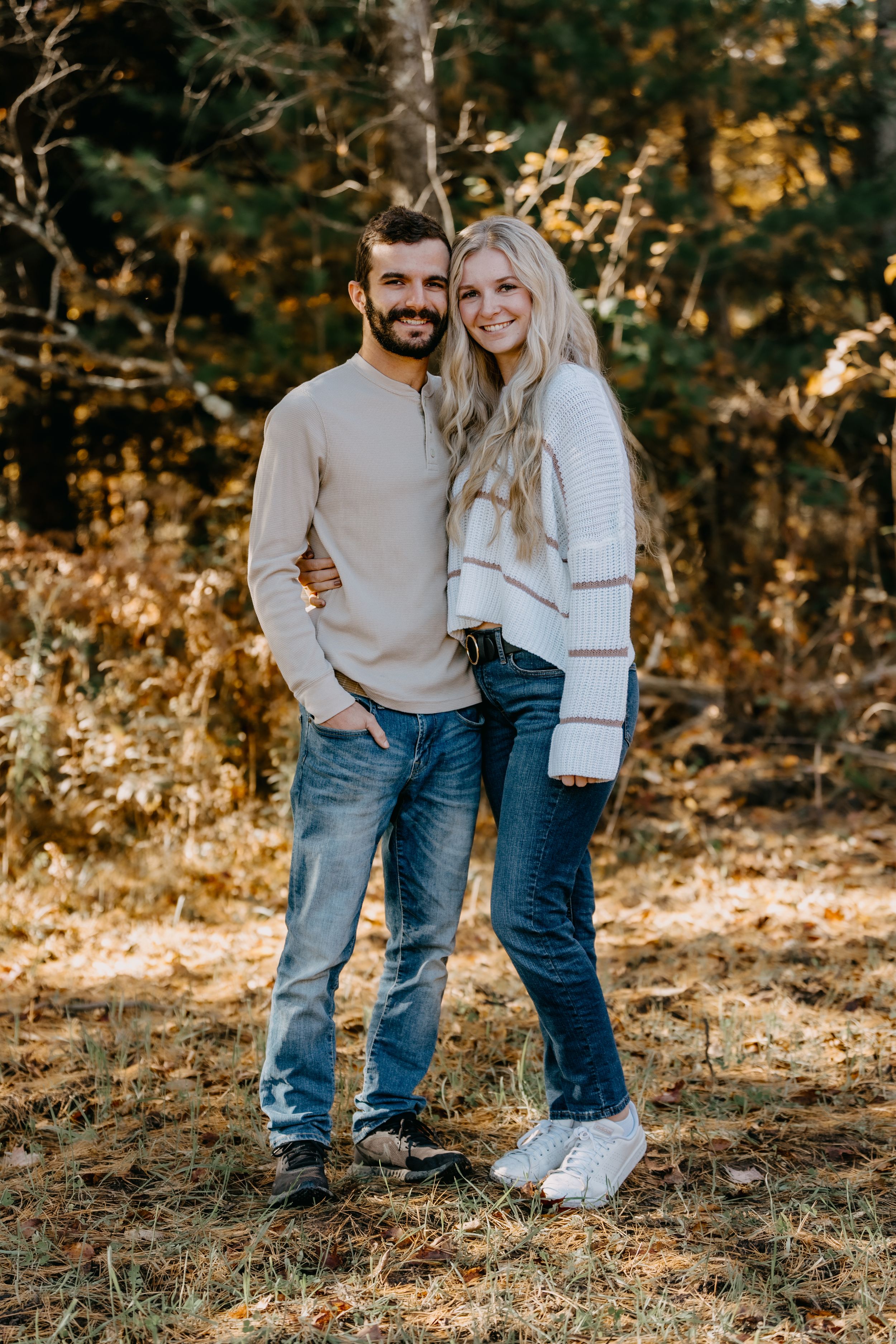 Couple wearing matching neutral sweaters and jeans stand together in autumn woodland setting with warm lighting.