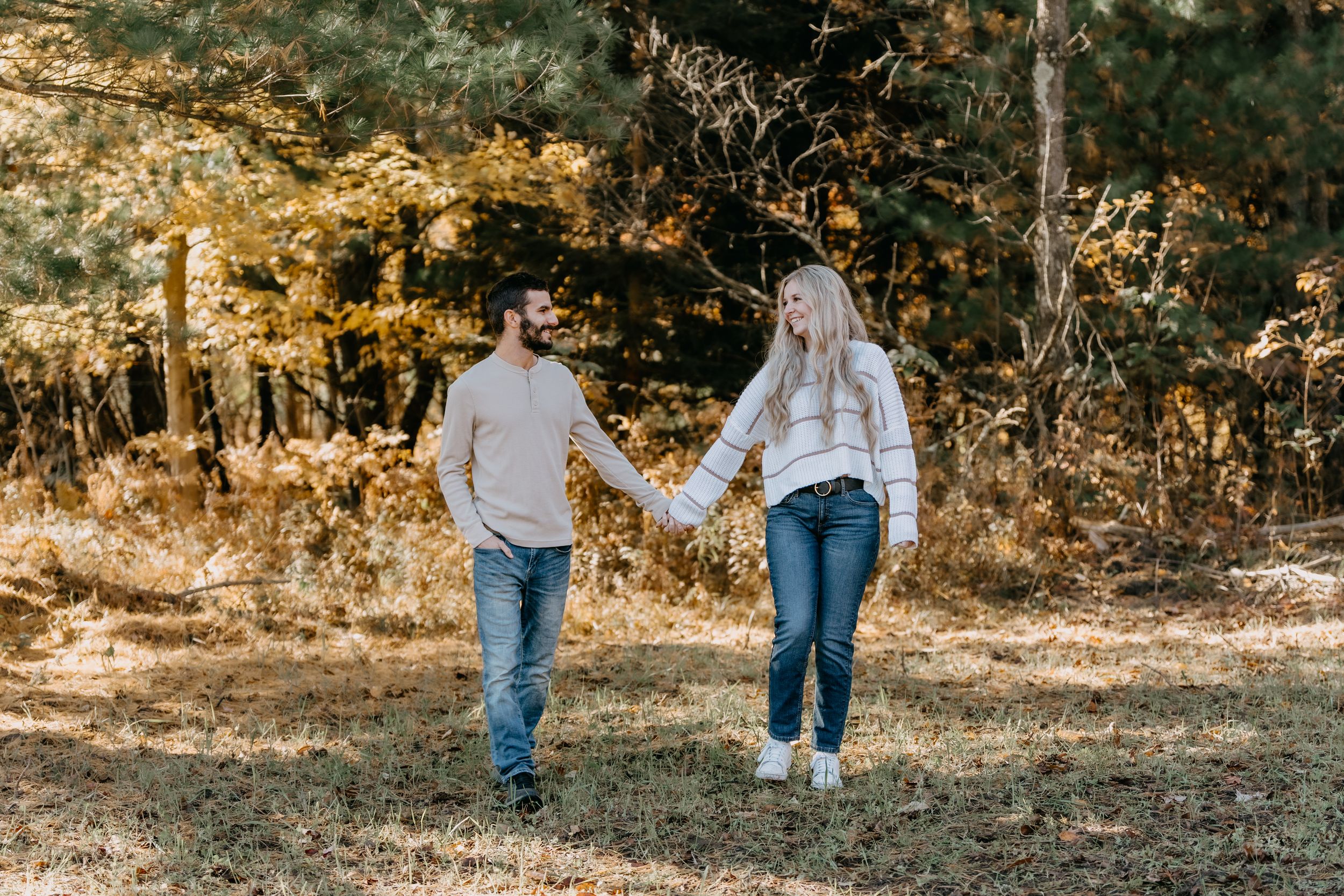 A couple holding hands while walking through a sunlit autumn forest with golden foliage in the background.