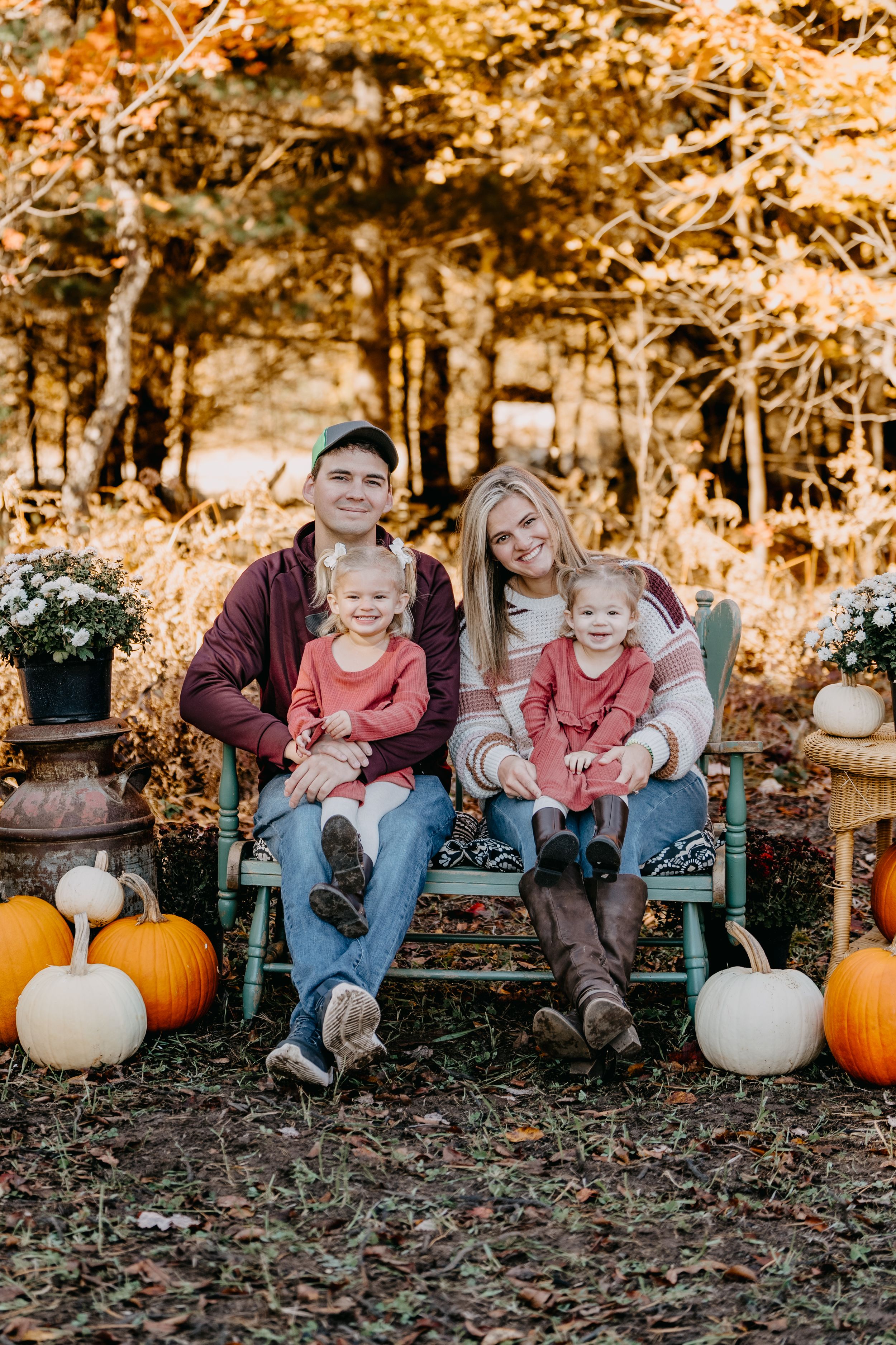 Family sits in an autumn scene among orange pumpkins and golden fall foliage on the forest ground.