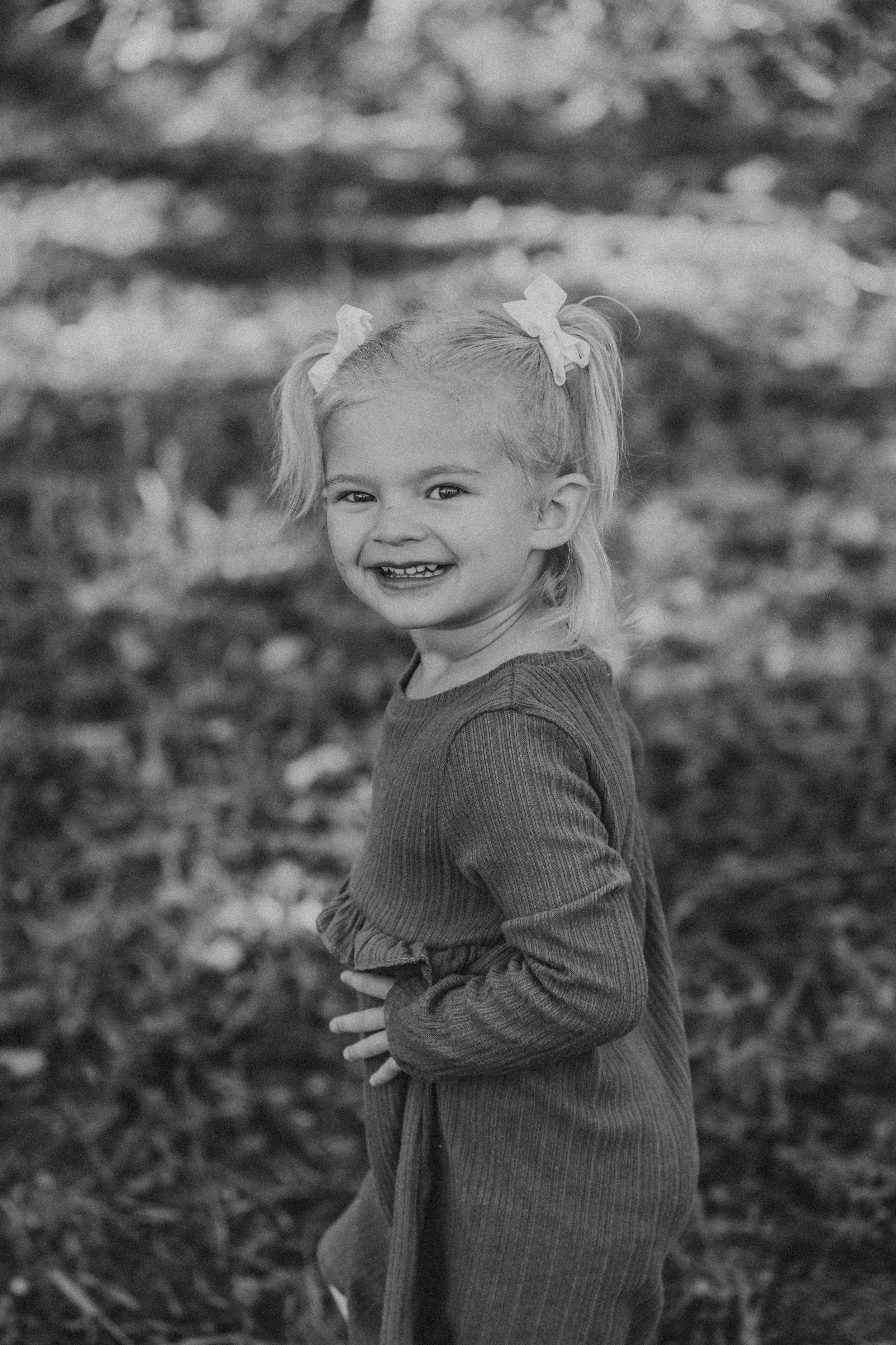 Black and white portrait of a young child with pigtails wearing a ribbed dress and smiling joyfully outdoors.