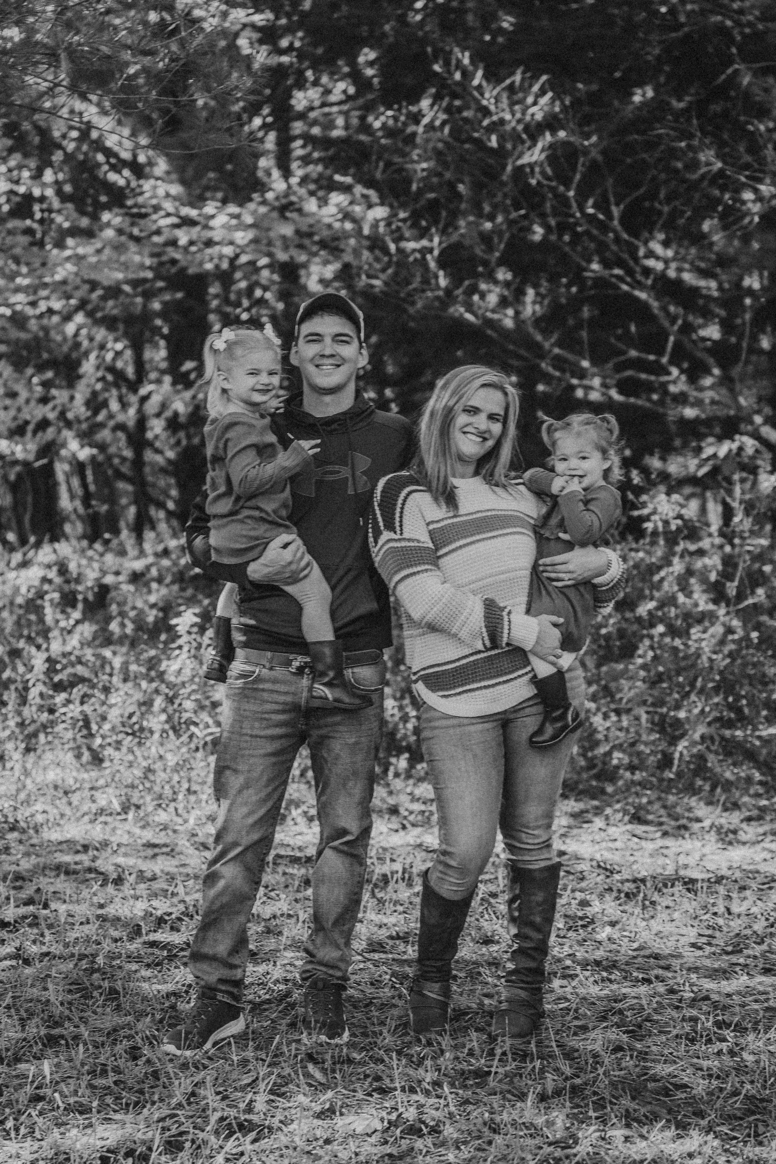 Young family stands together outdoors in a black and white photo with wooded background.
