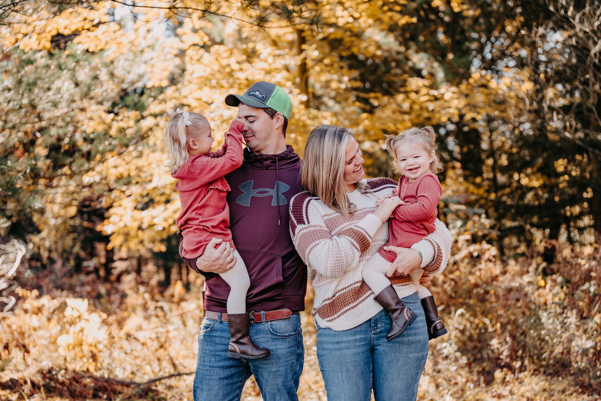 A family dressed in casual attire stands together outdoors during autumn with golden foliage in the background.