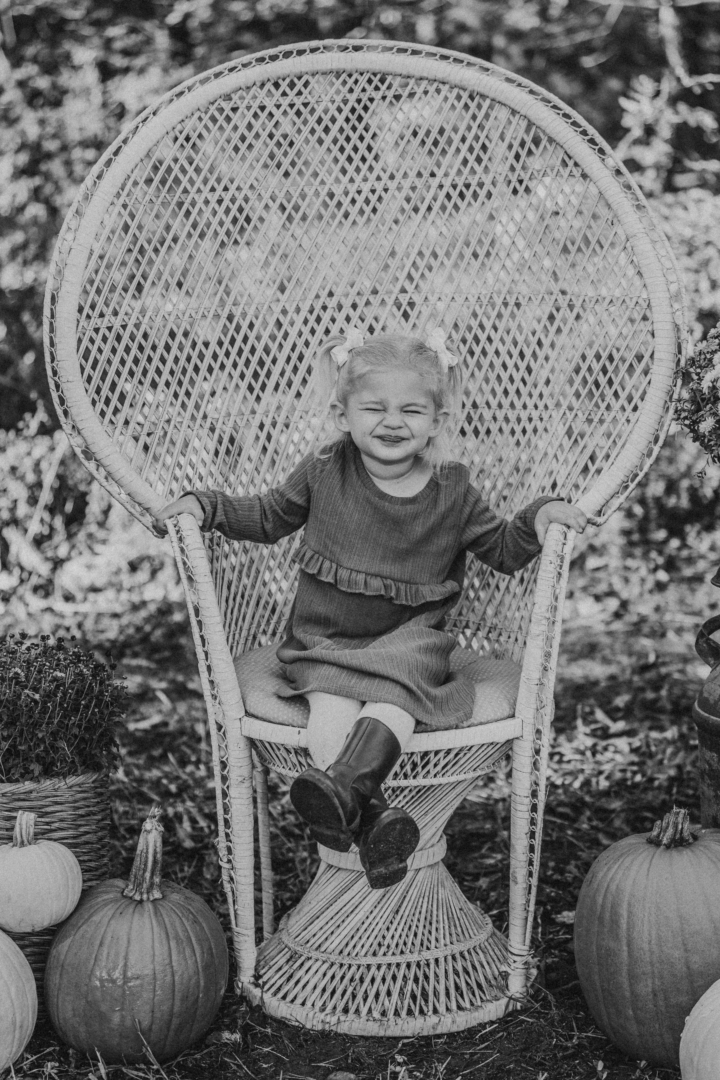 Child sits in large wicker peacock chair surrounded by autumn pumpkins in monochrome photo.