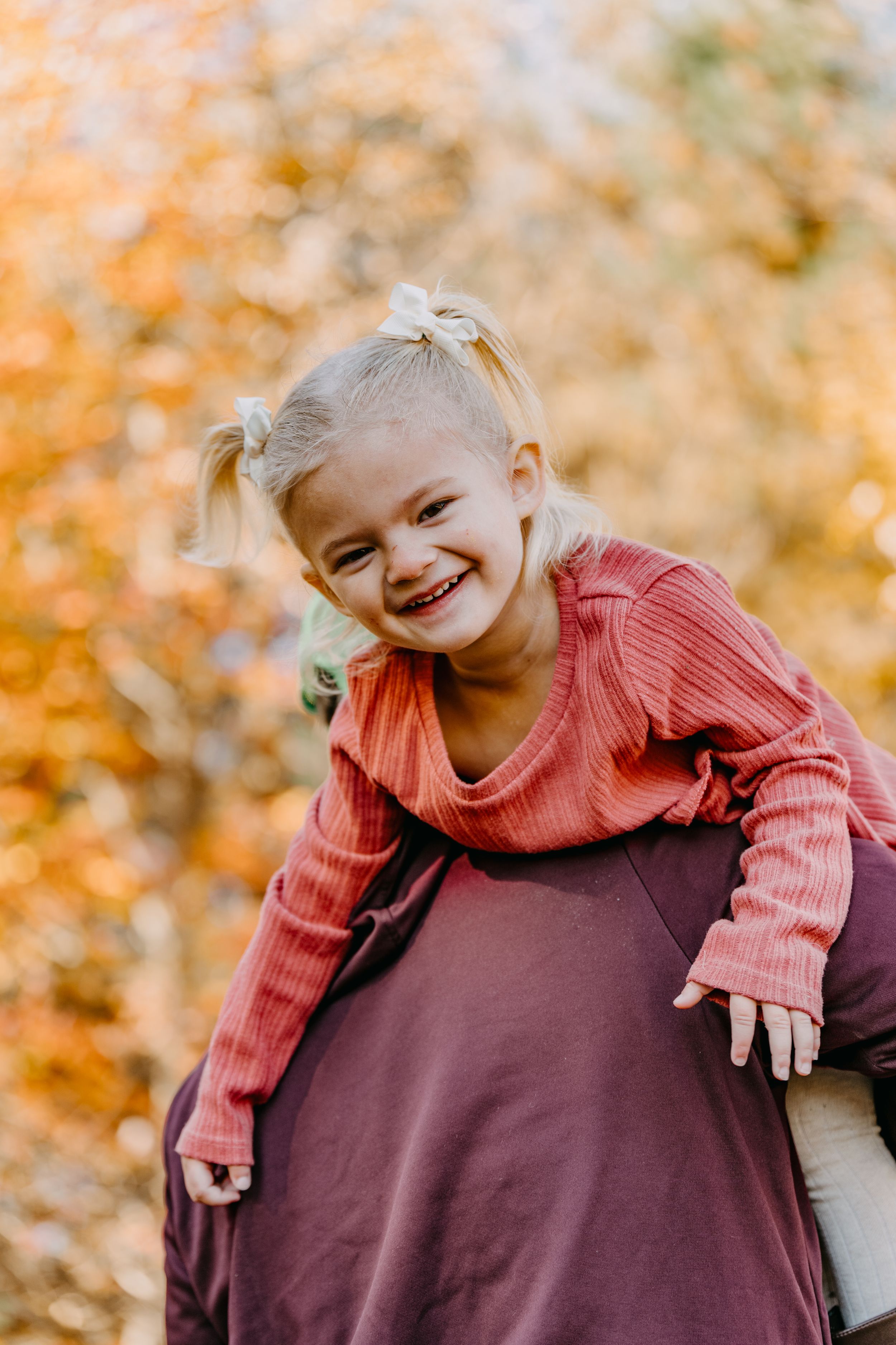 A smiling young child in a coral long-sleeve shirt poses playfully against an autumn backdrop with golden foliage.