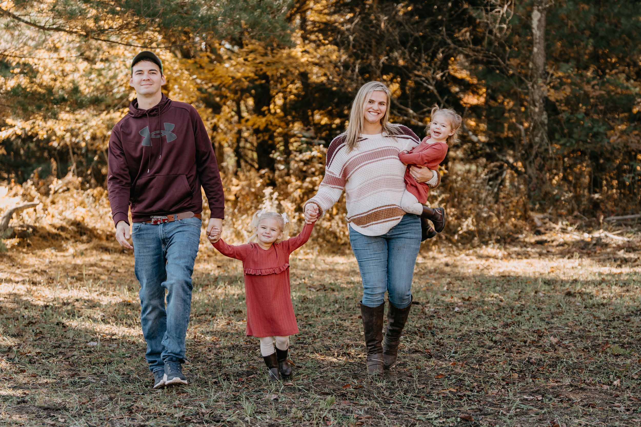 A family walks together holding hands through fallen autumn leaves in a wooded outdoor setting.