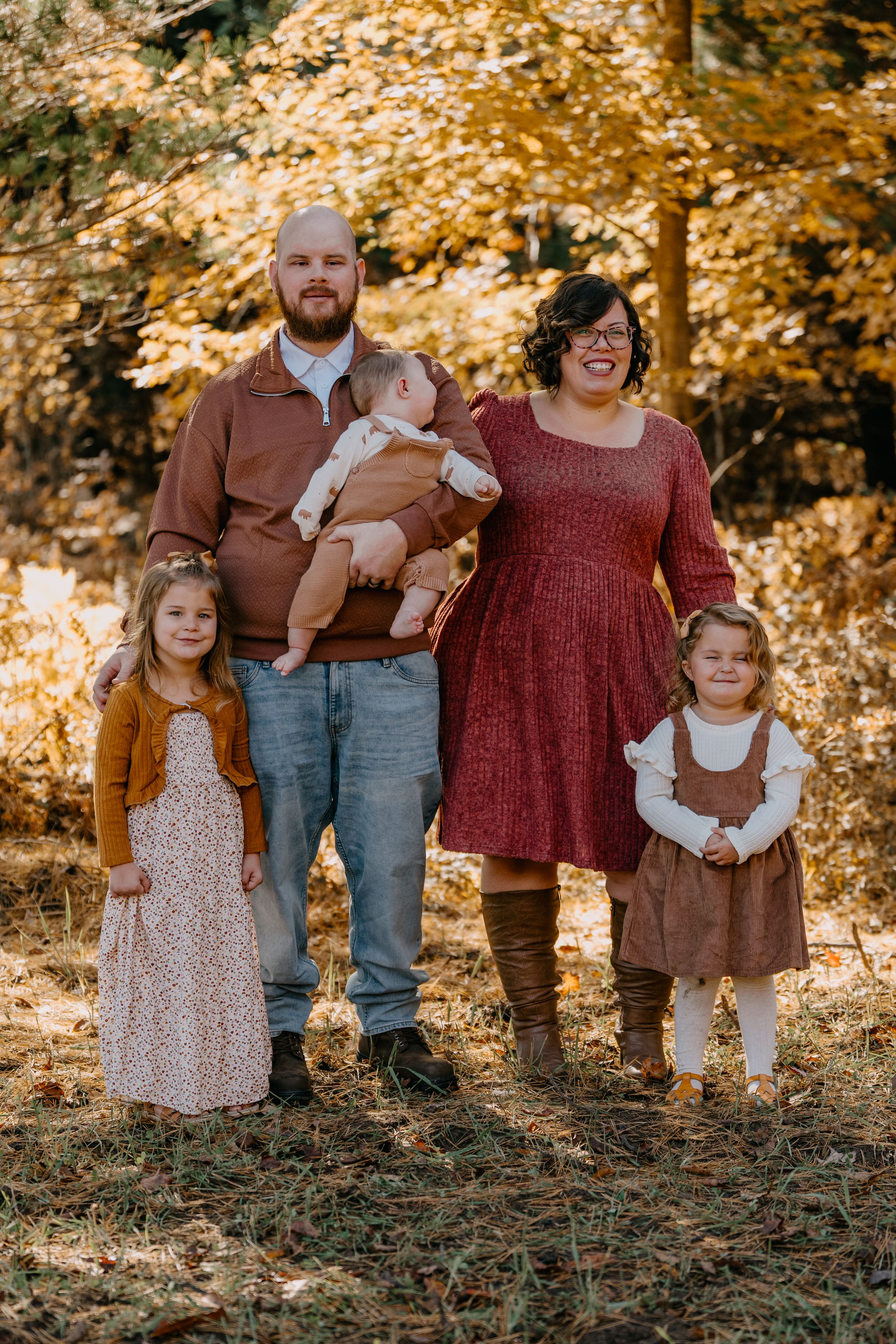 Family dressed in autumn colors poses together in front of golden fall foliage on a sunny day.