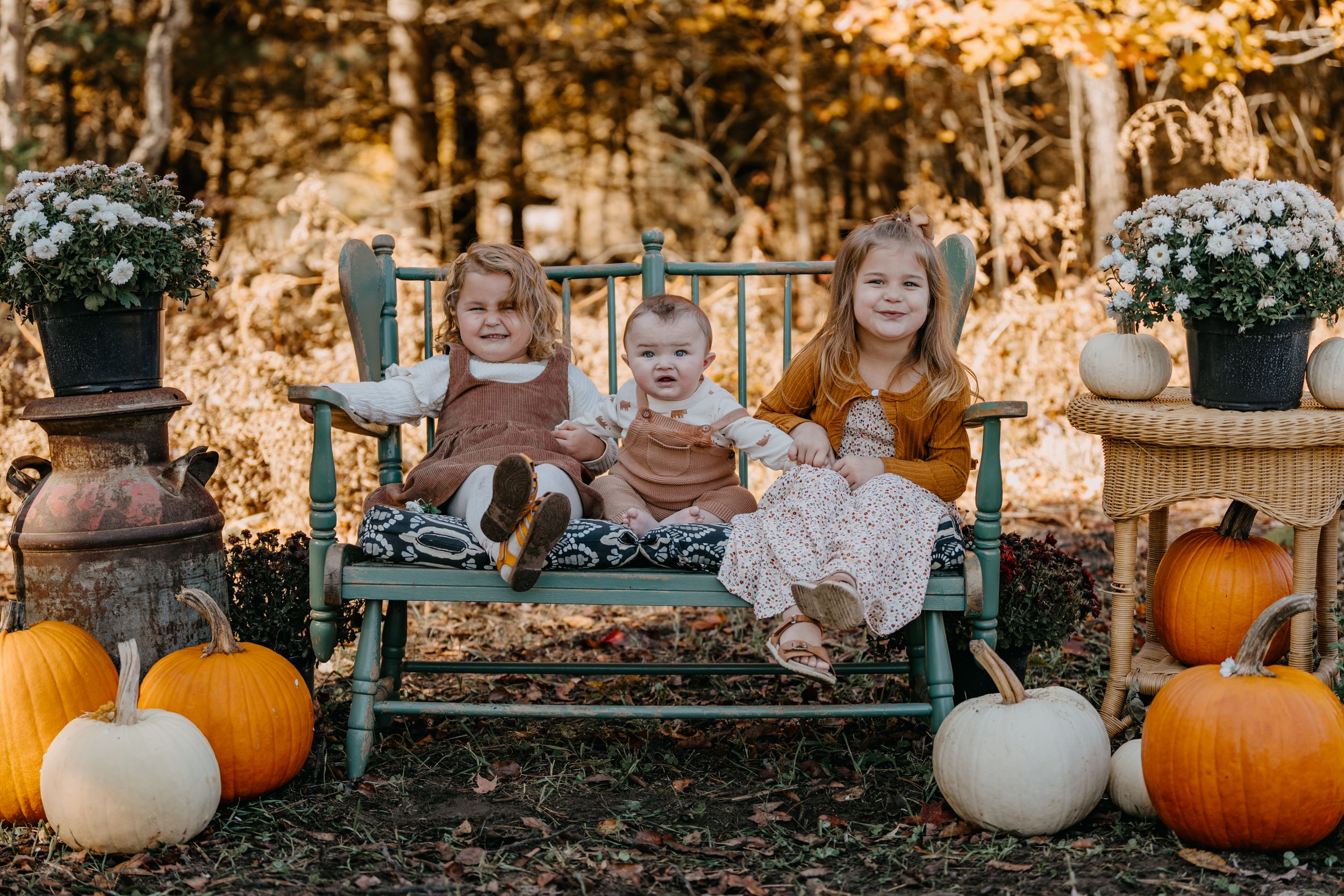 Fall harvest scene with children sitting on rustic bench surrounded by orange pumpkins and fall flowers.