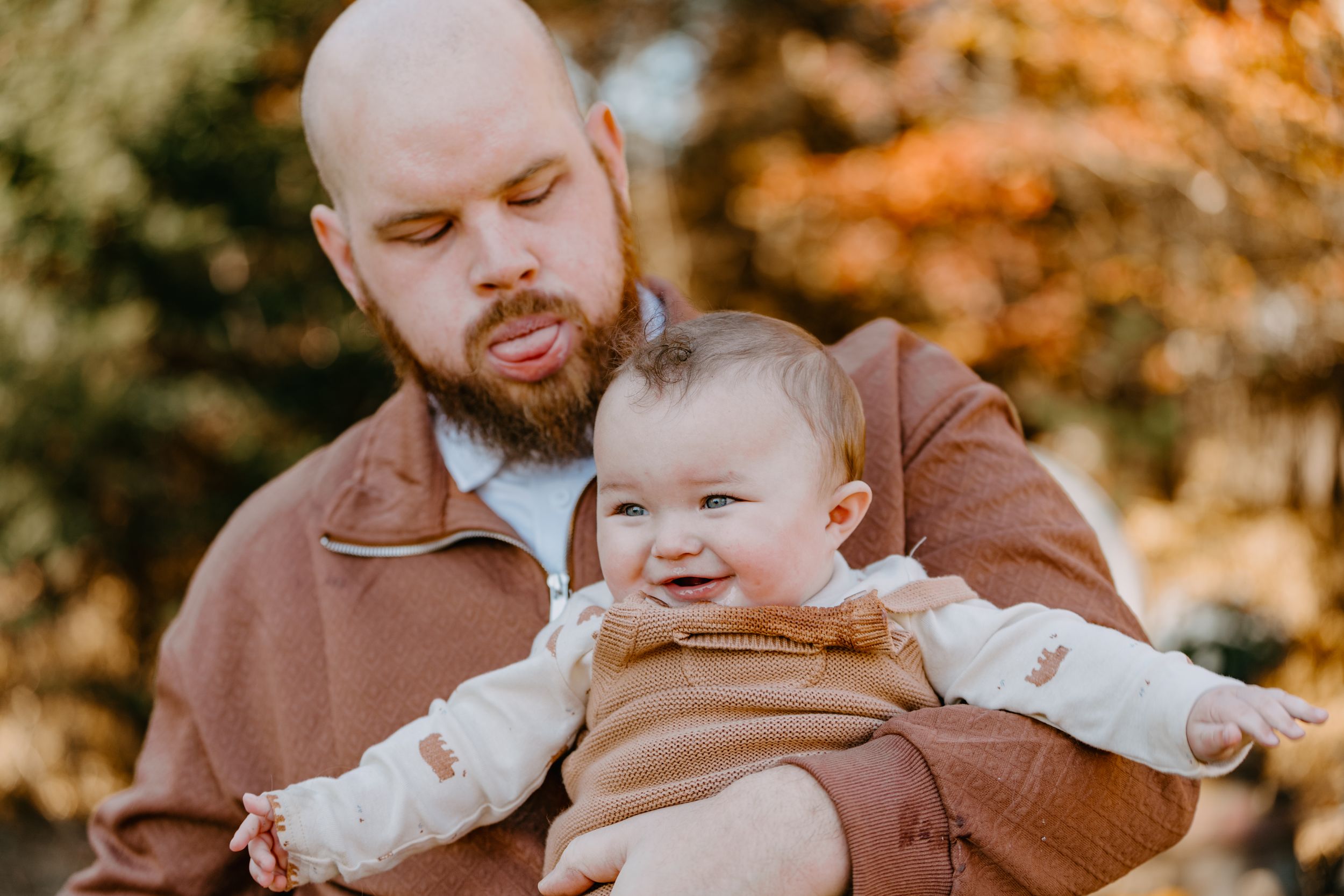 Parent holds a smiling baby wearing a brown outfit during an autumn outdoor photo session with warm golden lighting.