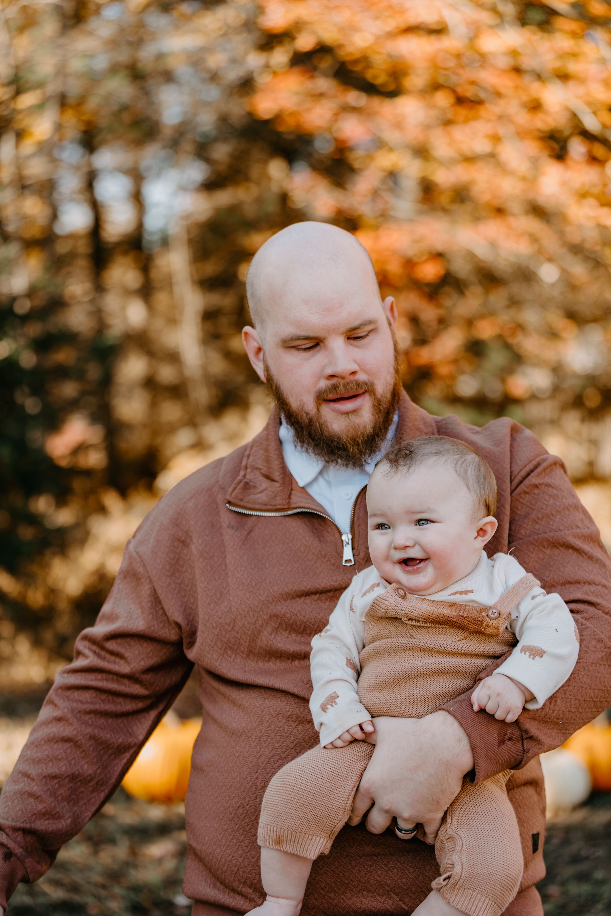 A heartwarming outdoor autumn scene shows a bearded person in brown jacket holding a joyfully smiling baby in matching attire.