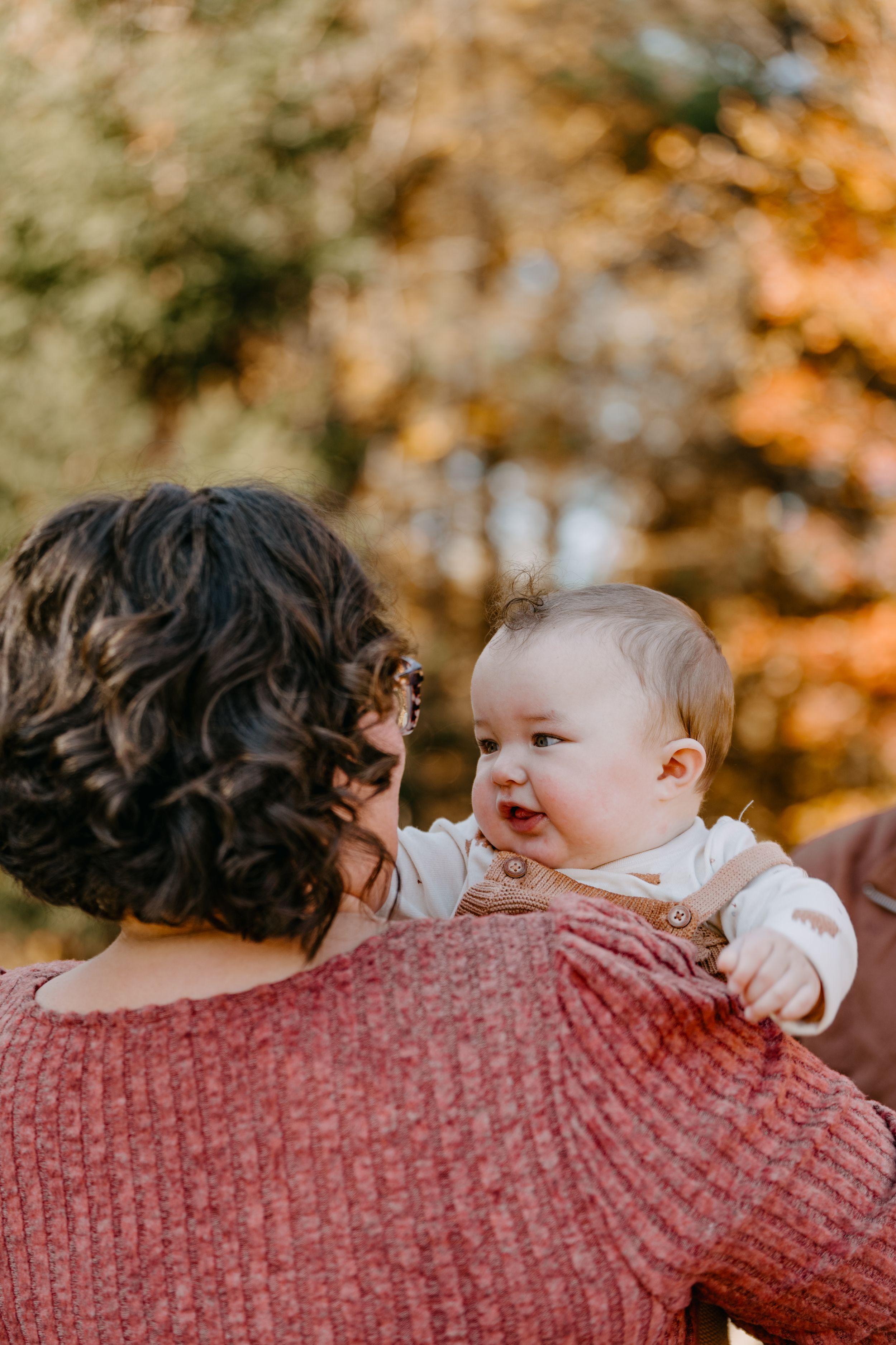 A smiling baby in a white sweater rests on someone's shoulder during a warm autumn day with golden foliage in background.