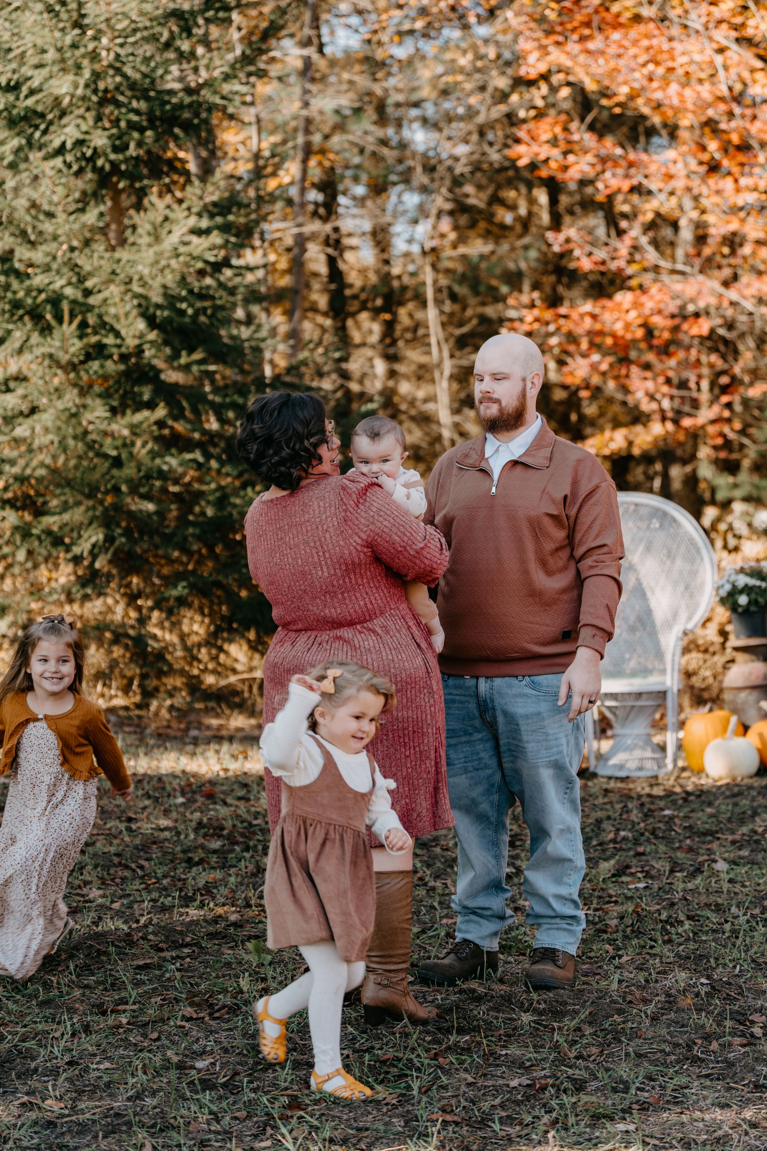 Family stands together outdoors during autumn photoshoot with pumpkins and fall foliage in the background.
