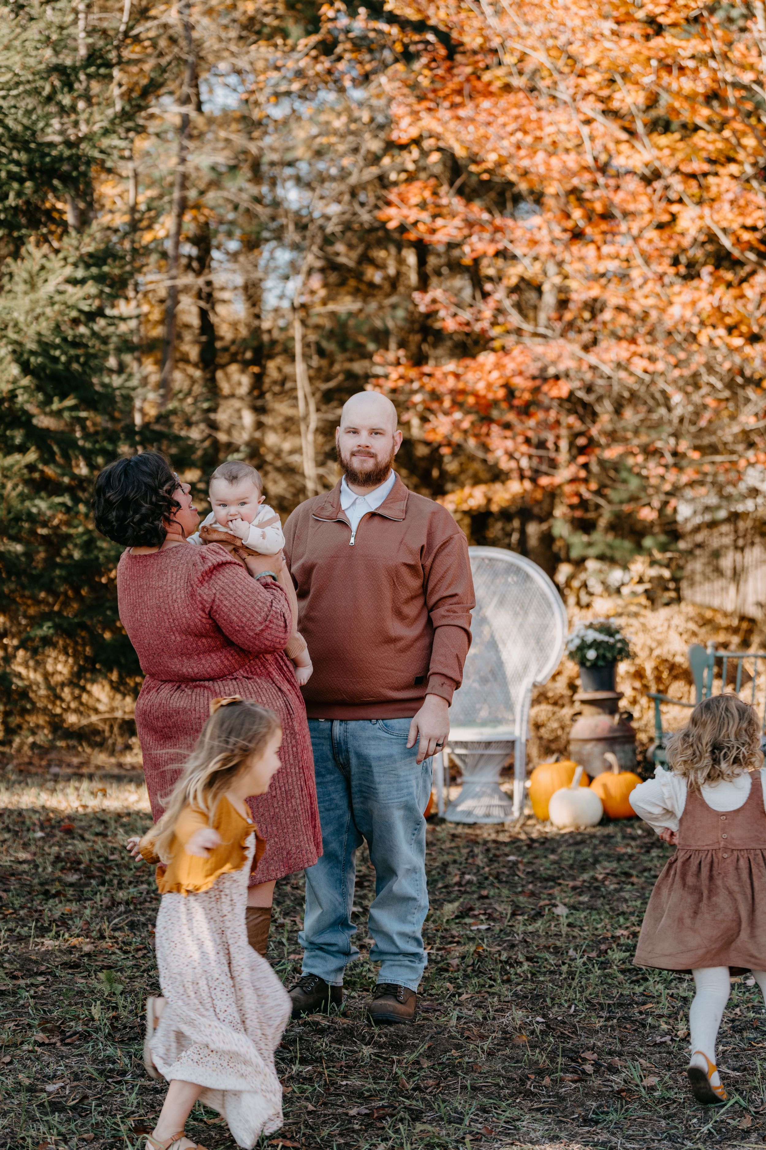 A fall scene with a family outdoors surrounded by autumn foliage and decorative pumpkins on the ground.