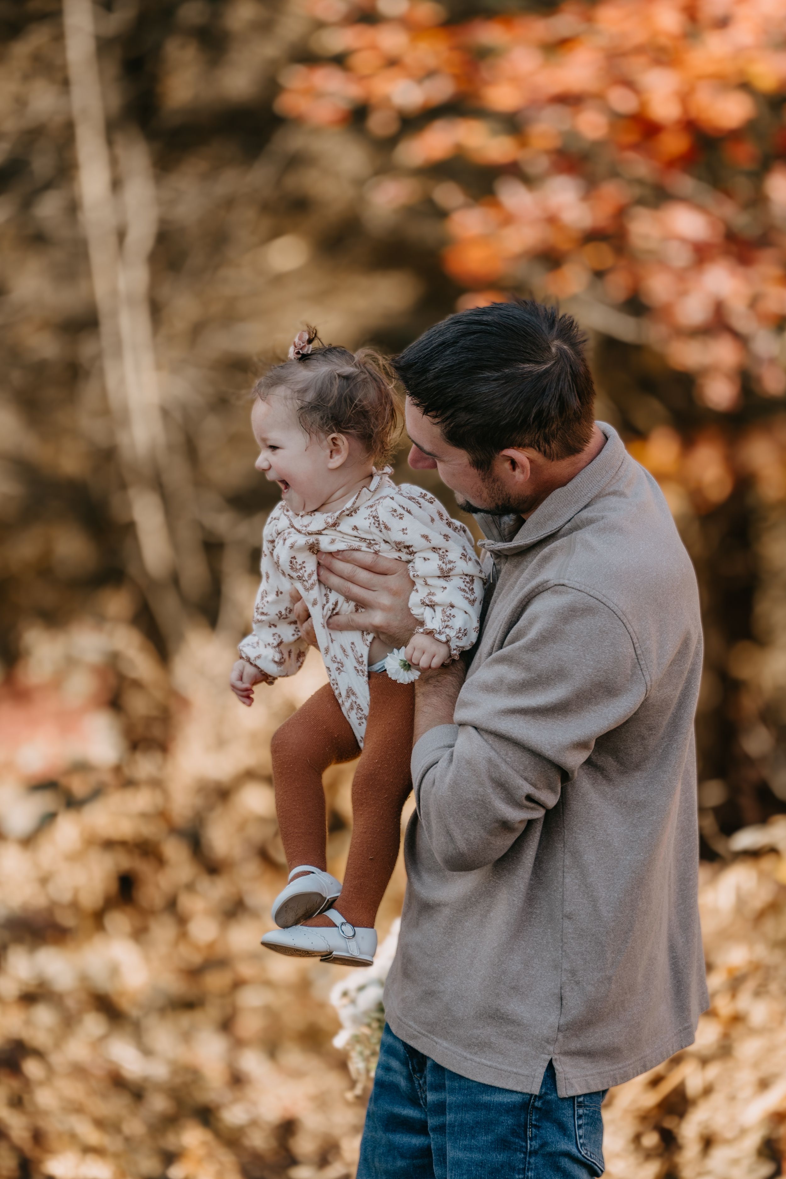 A parent holds and plays with a laughing child outdoors during autumn with orange foliage in the background.