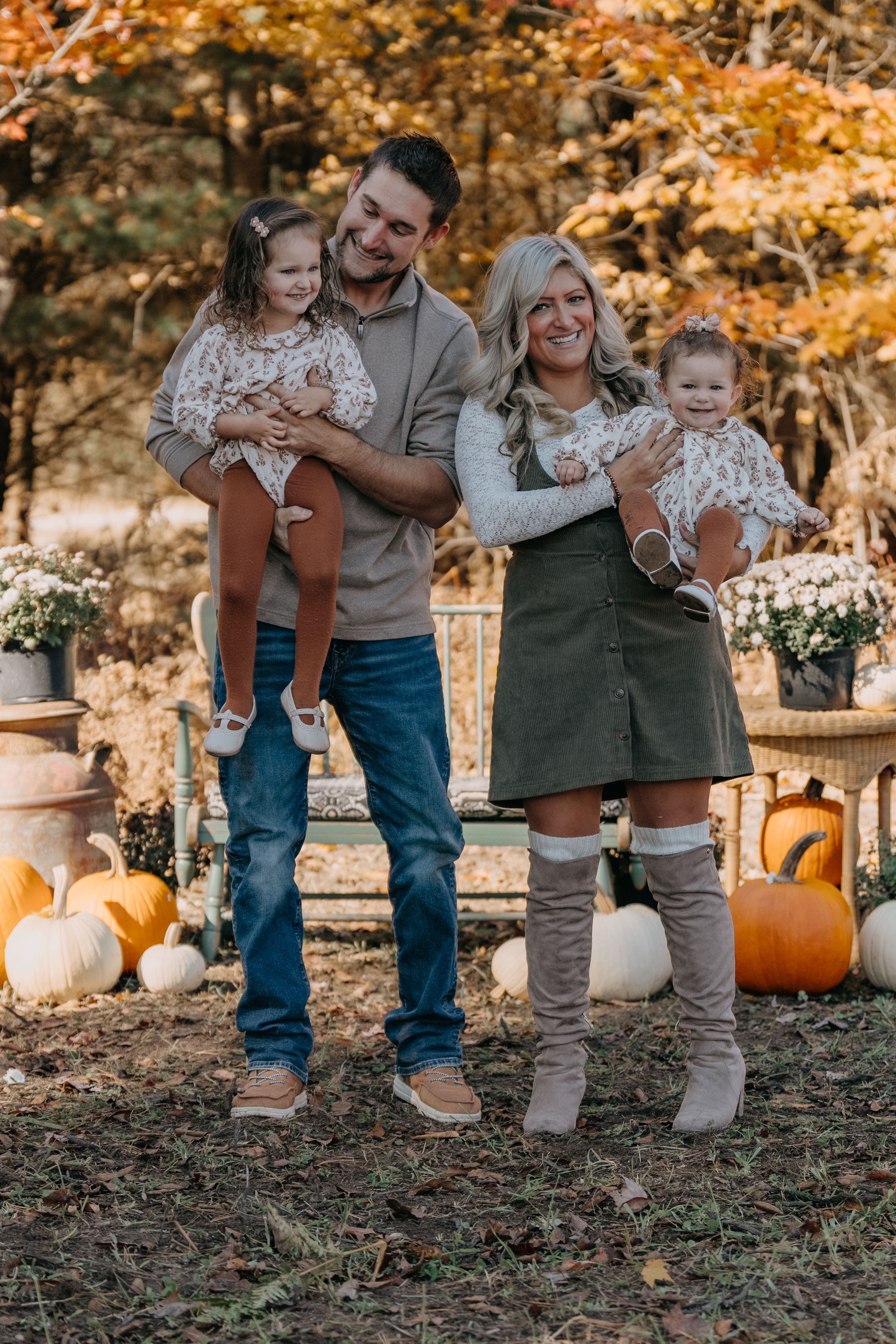 A family poses for a fall portrait outdoors with pumpkins and autumn foliage in warm earth tones.