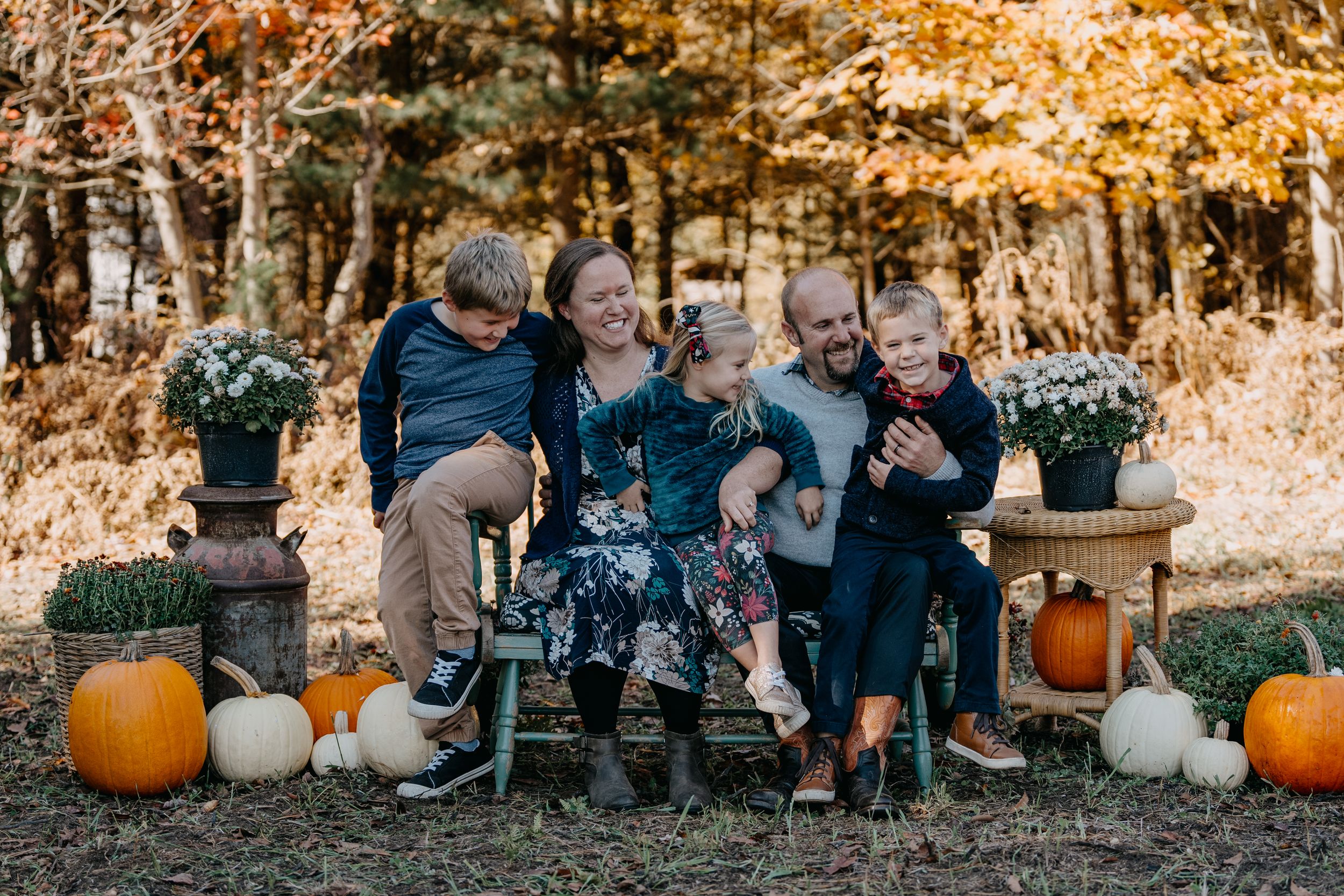 Family sits on fallen log surrounded by autumn pumpkins and colorful foliage during outdoor photo session.
