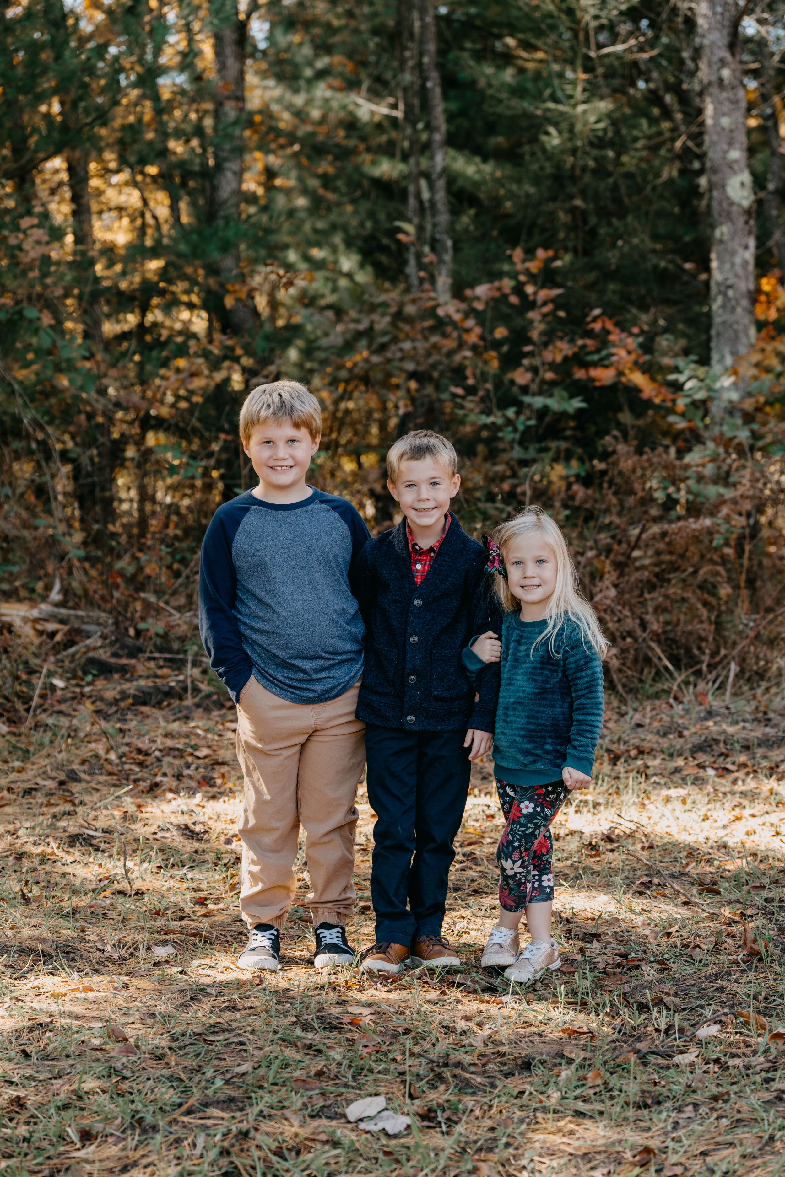 Three children stand together on a forest path during autumn with fallen leaves and evergreen trees in the background.