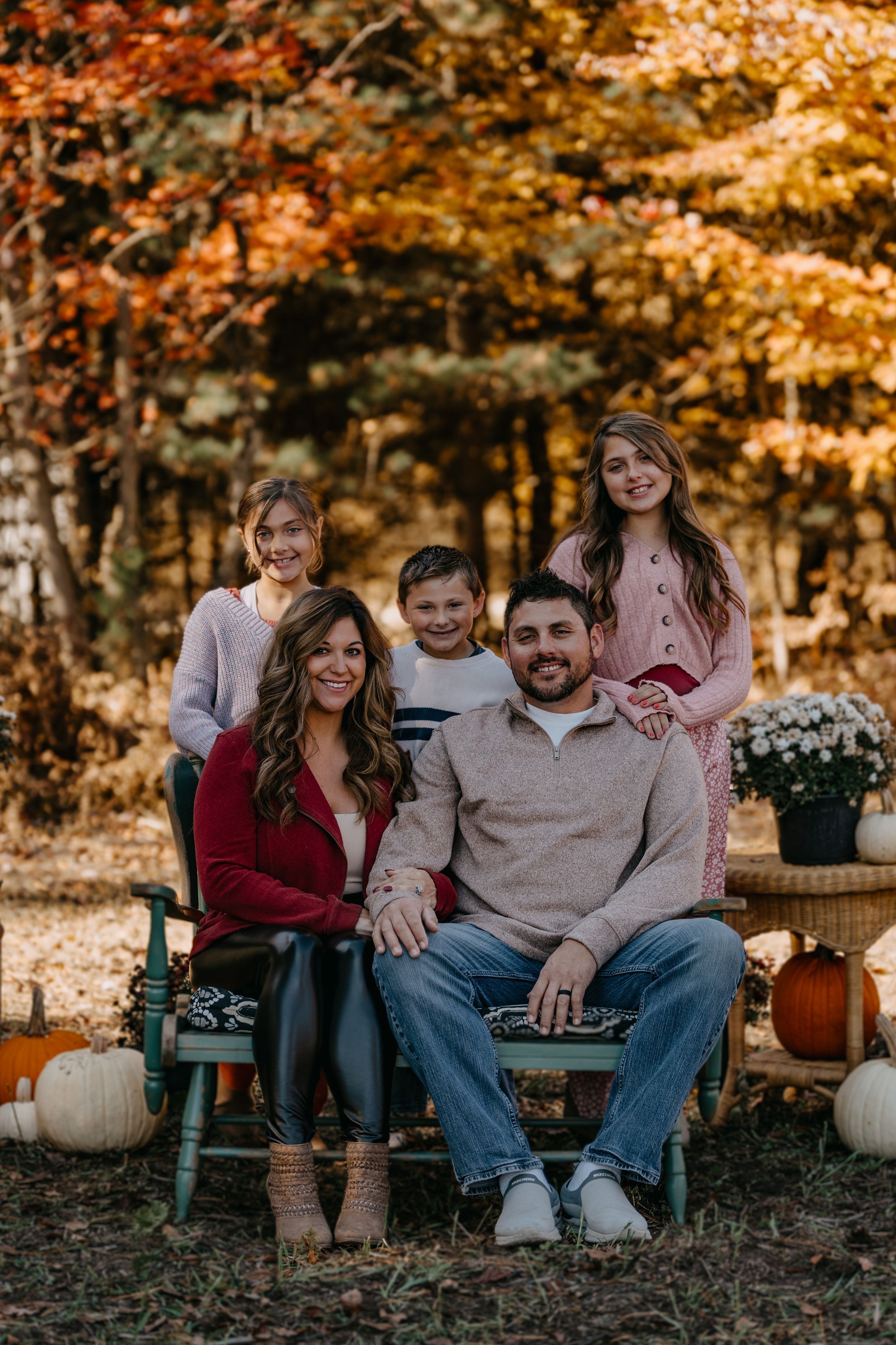 A family sits together on outdoor chairs surrounded by fall foliage and autumn decorations in warm golden lighting.