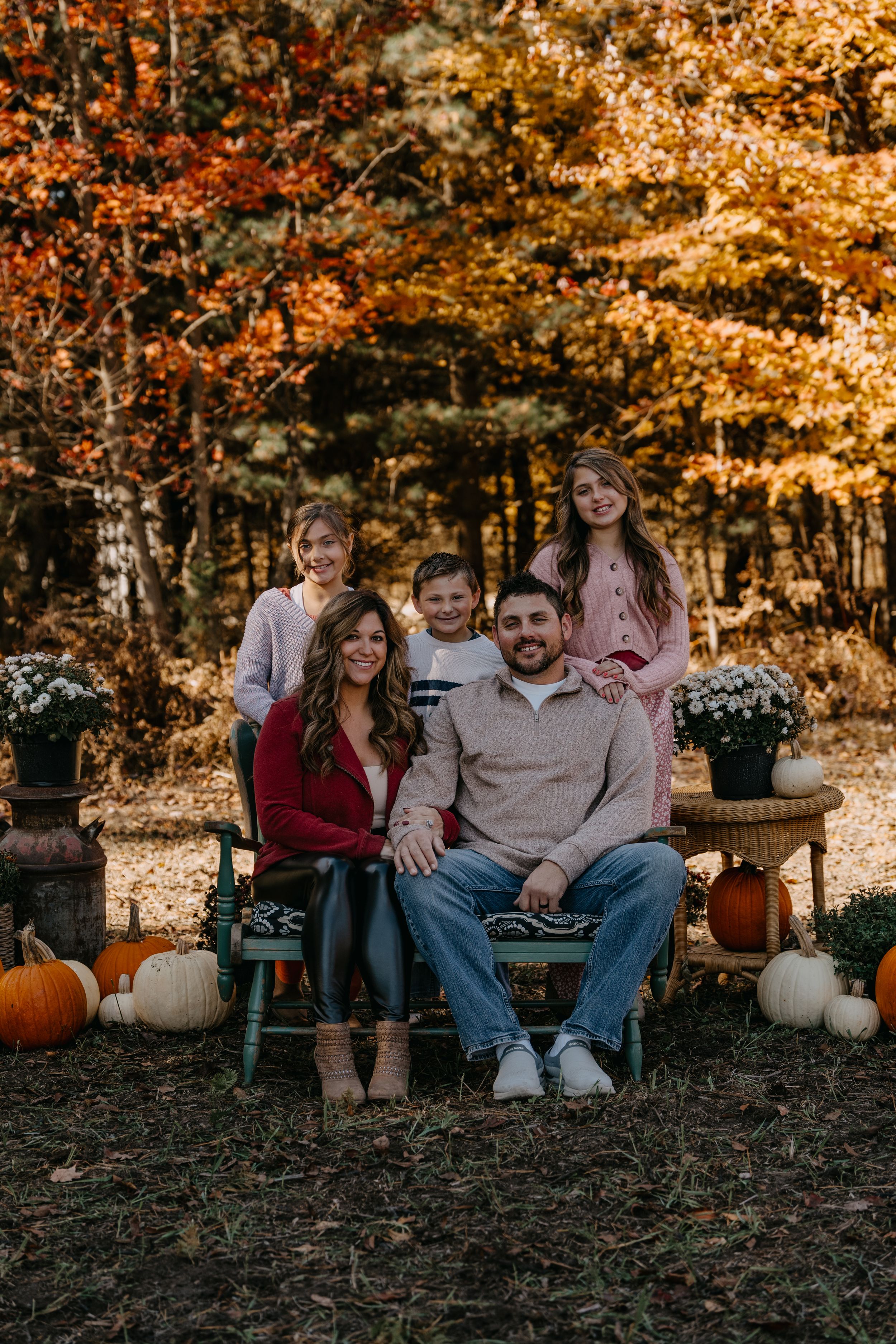 Group sitting on bench surrounded by autumn trees, pumpkins and fall decor in rustic outdoor setting.
