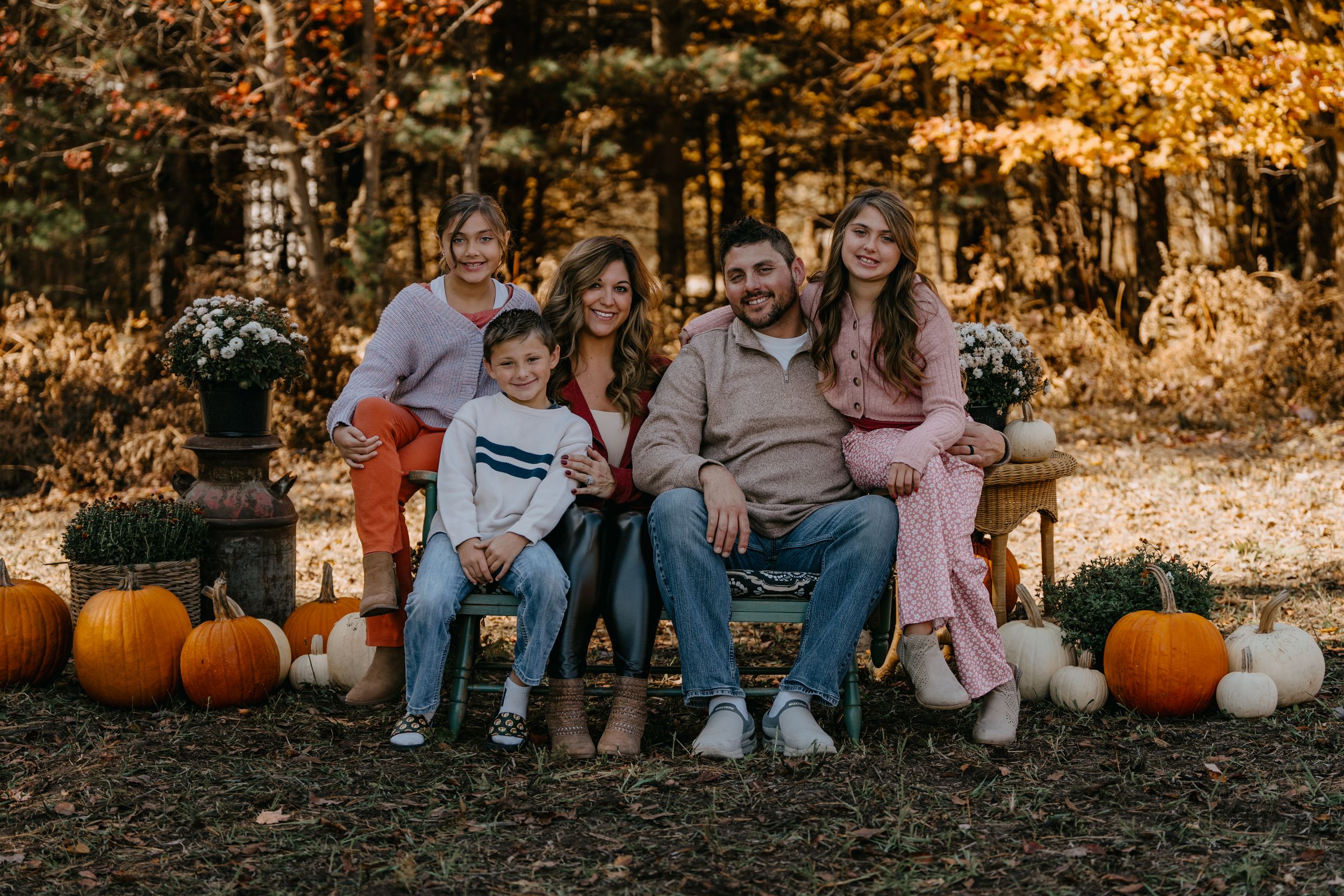 Group sitting on bench among autumn pumpkins and golden fall foliage in outdoor setting at sunset.