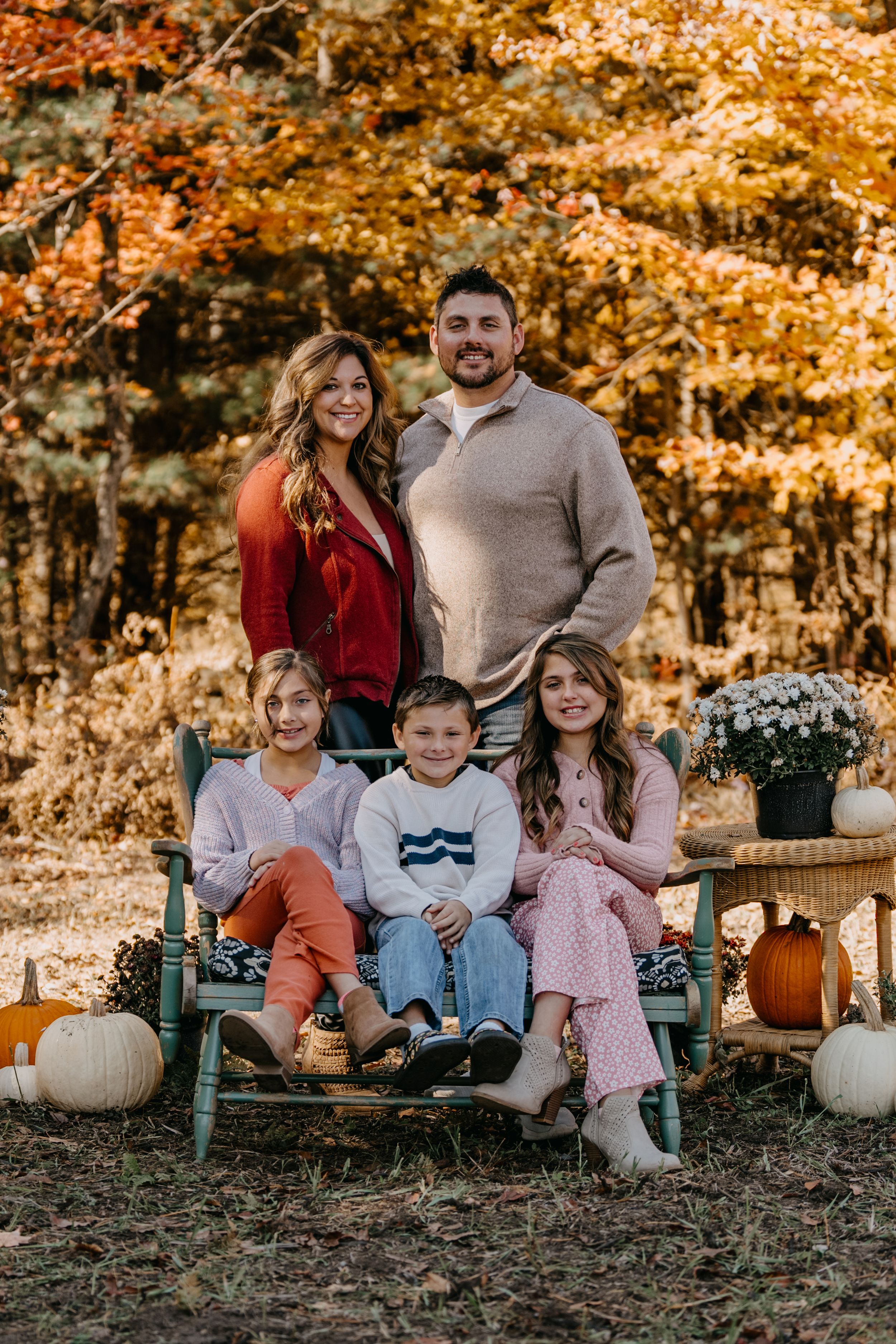 Family of five poses on rustic bench surrounded by fall pumpkins and autumn foliage in outdoor setting.
