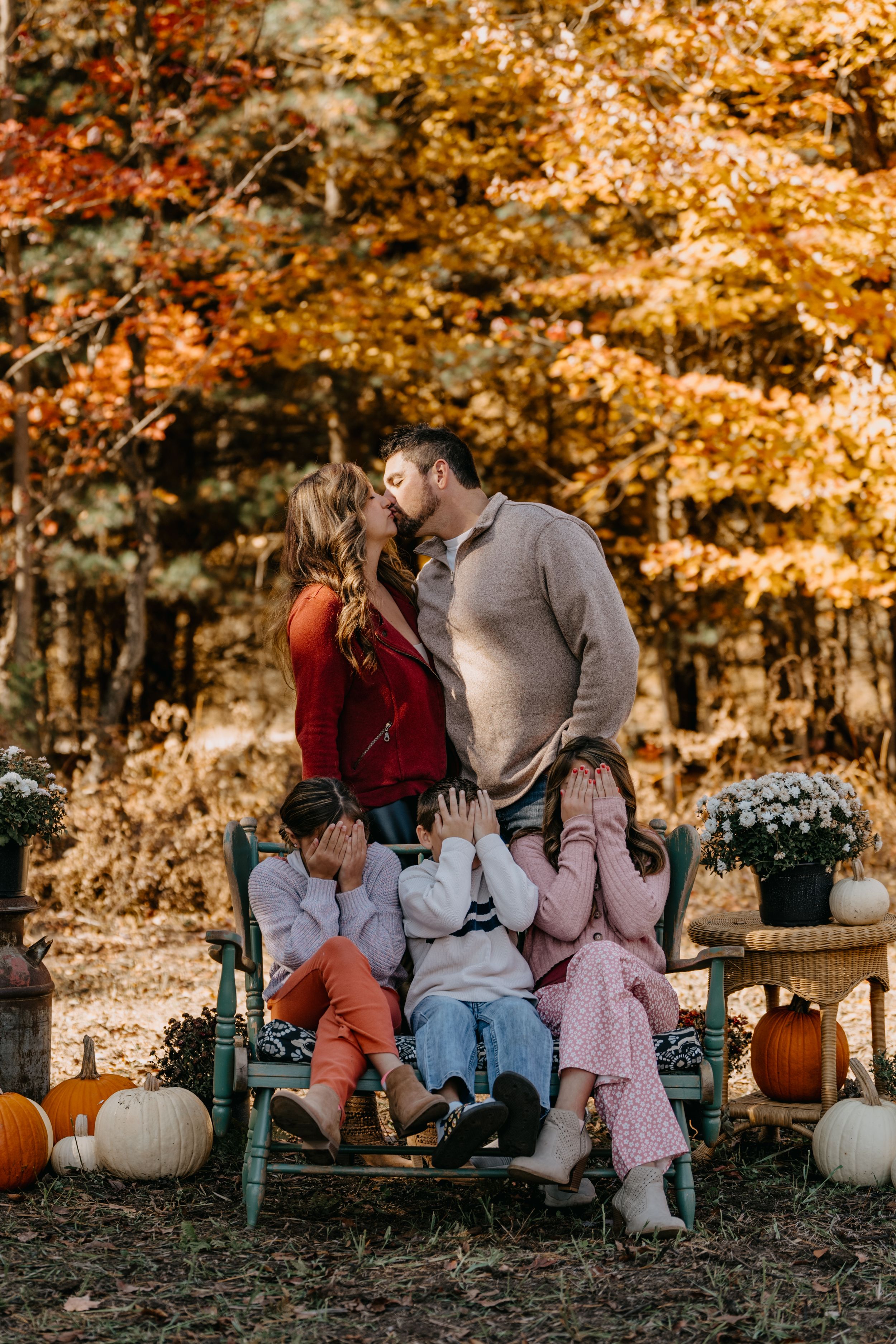 Fall family photo scene with pumpkins and mums featuring group posed on vintage bench against autumn foliage background.