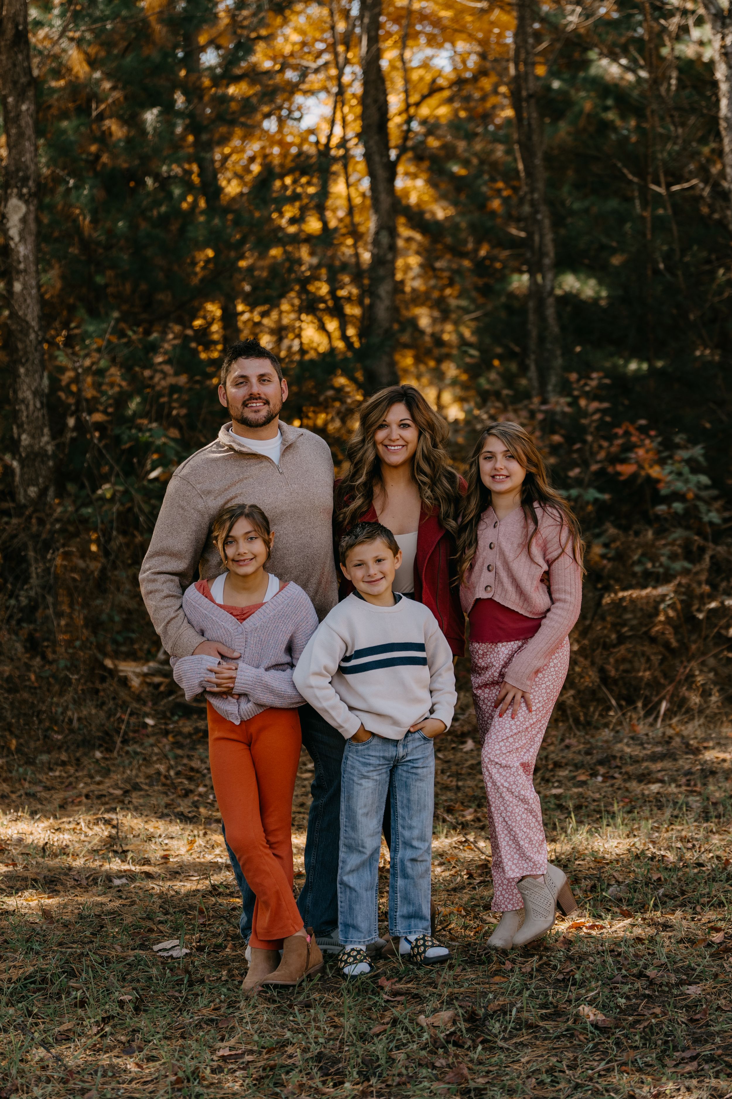 A family poses together in casual fall attire against a backdrop of autumn trees with golden leaves at sunset.