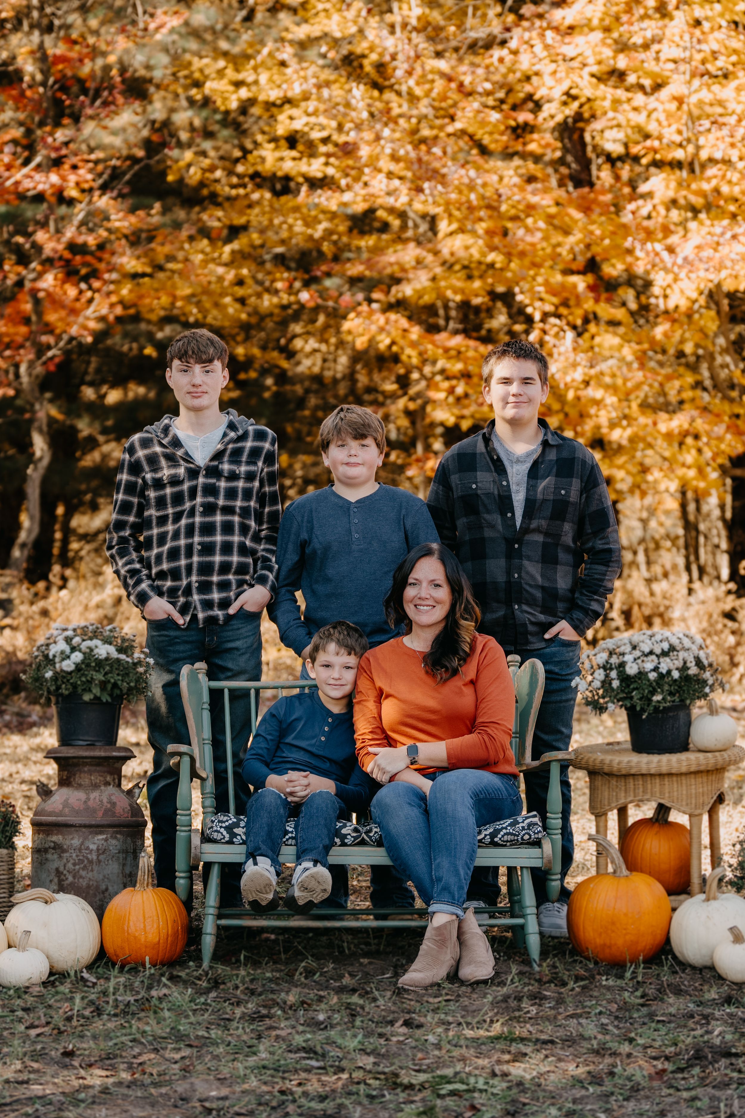 Family poses on rustic bench with autumn foliage and decorative pumpkins in seasonal outdoor setting.