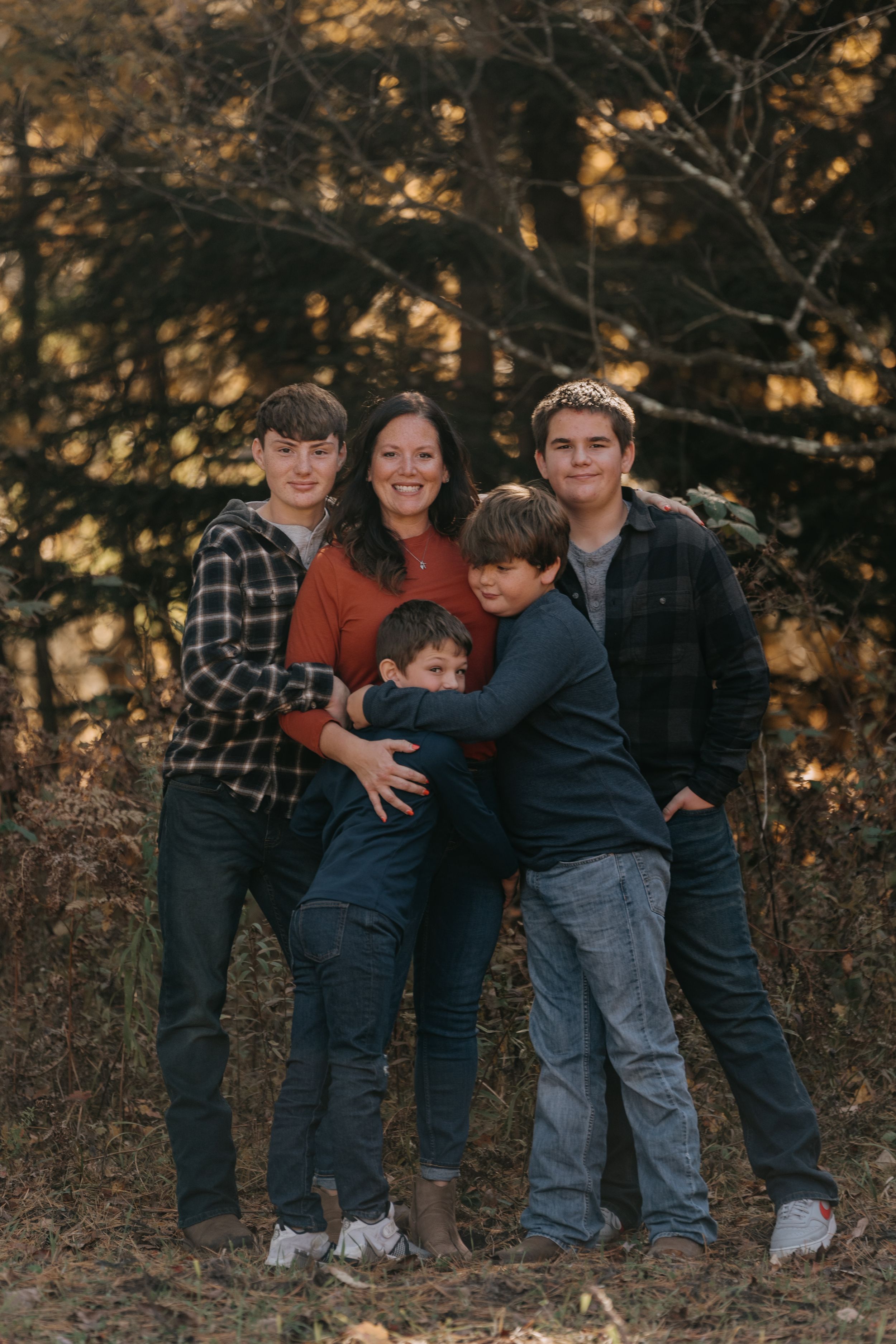 A group shares warm embraces and laughter during an outdoor autumn family photo session near trees and fallen leaves.