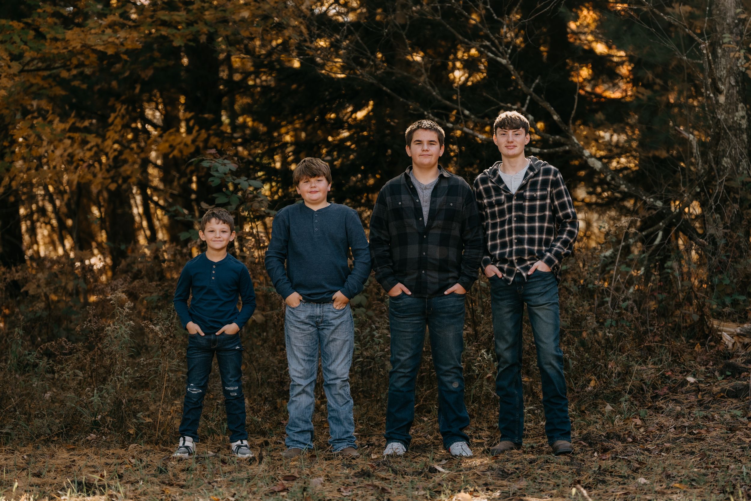 Four people stand together outdoors in casual attire against an autumn forest backdrop with golden foliage.