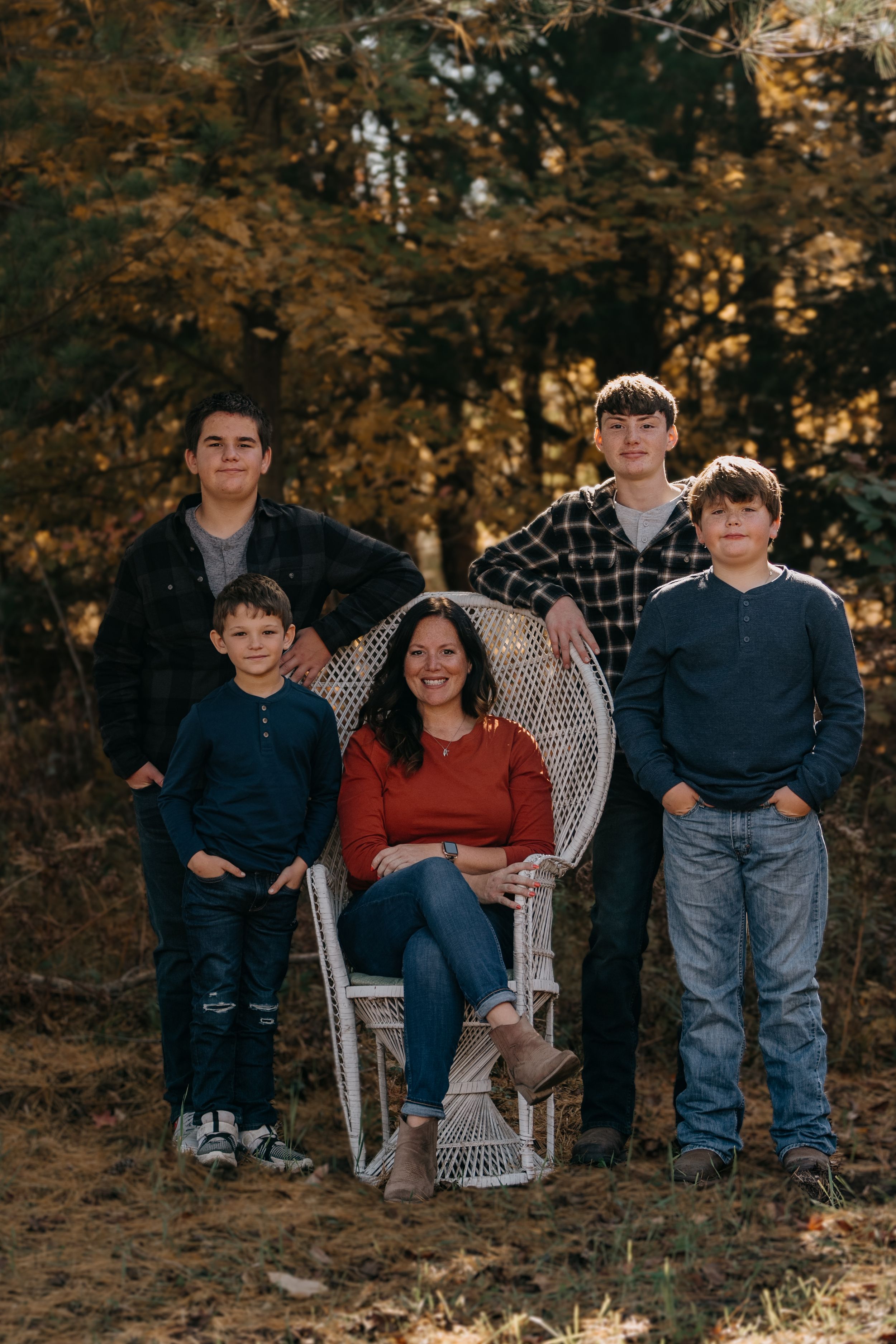 A group in casual fall attire poses outdoors against autumn foliage, with a wicker chair as the central focal point.