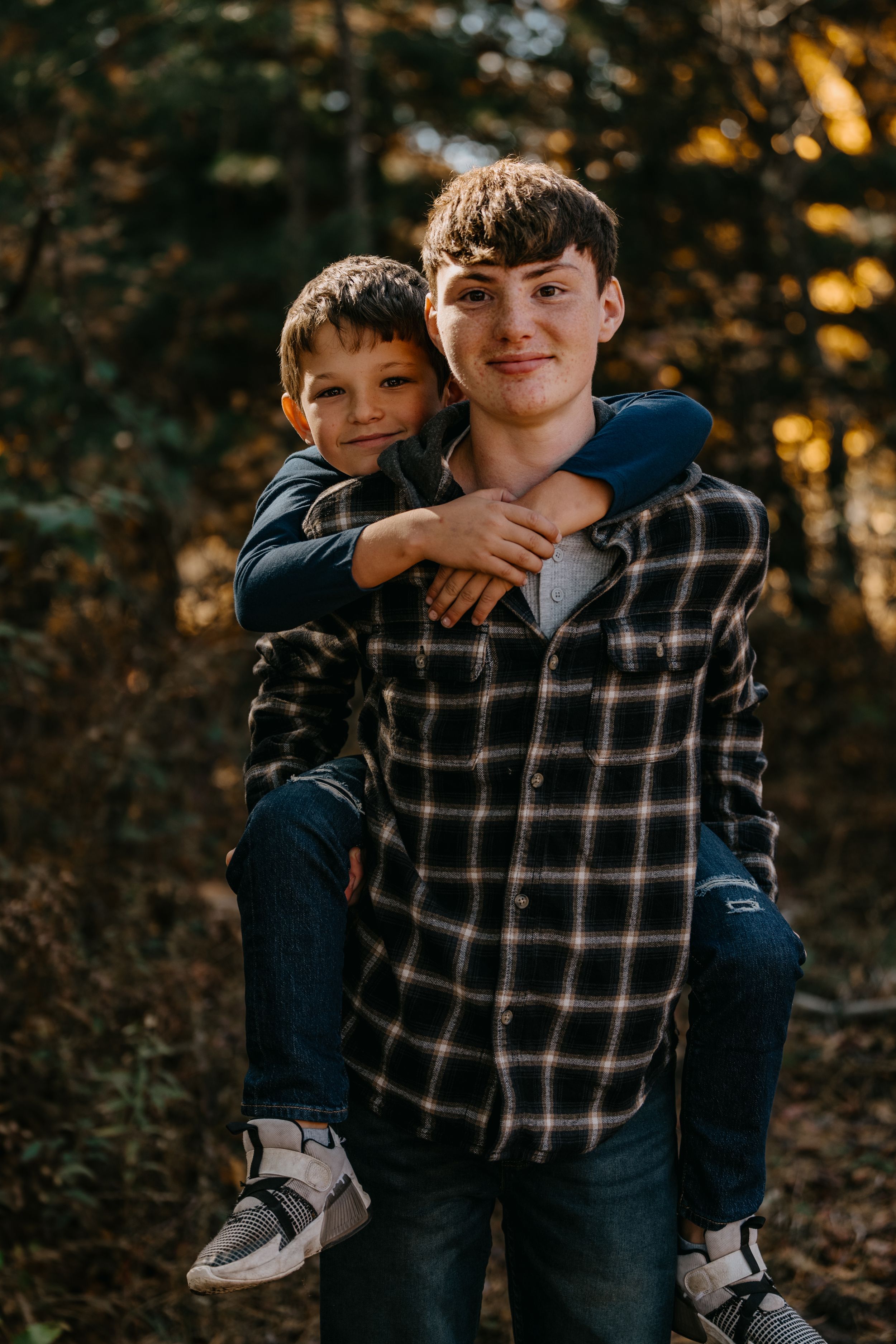 A young person carrying another on their back while standing in a forest during golden hour wearing plaid flannel.