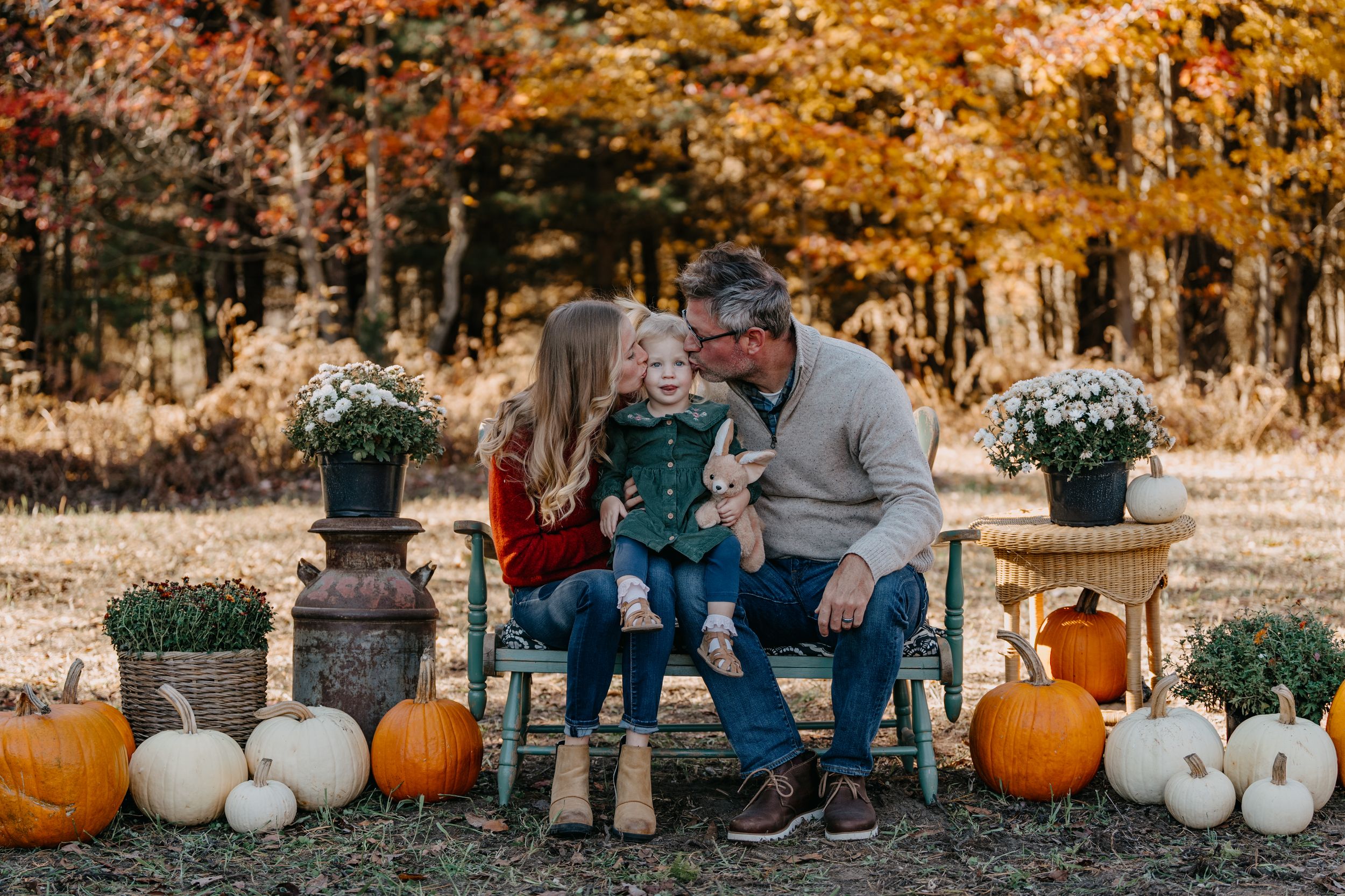 Three people sit on a rustic bench surrounded by autumn decor including pumpkins and potted mums in a fall forest setting.