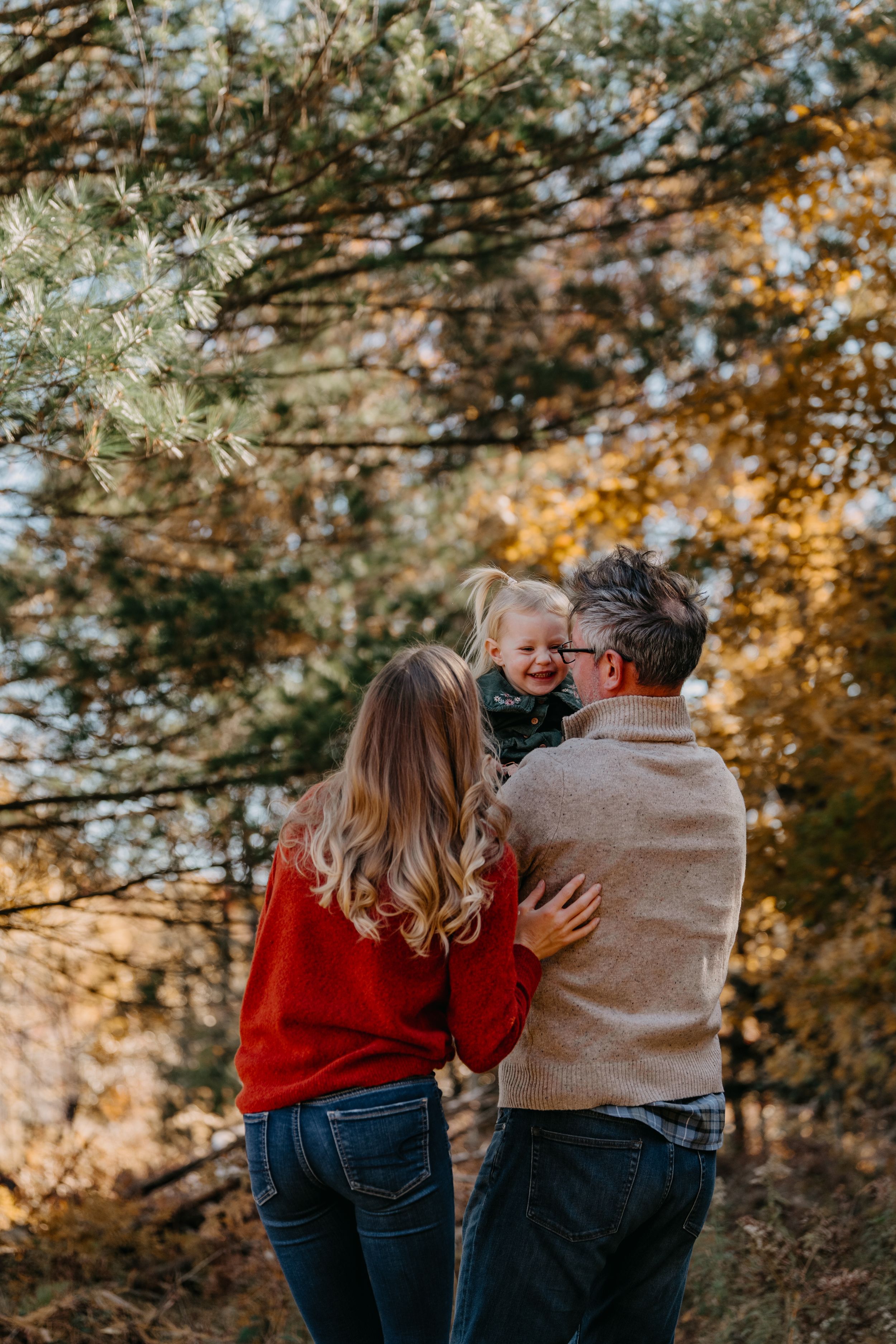Couple wearing red and beige sweaters embrace while walking through a fall forest setting during golden hour.