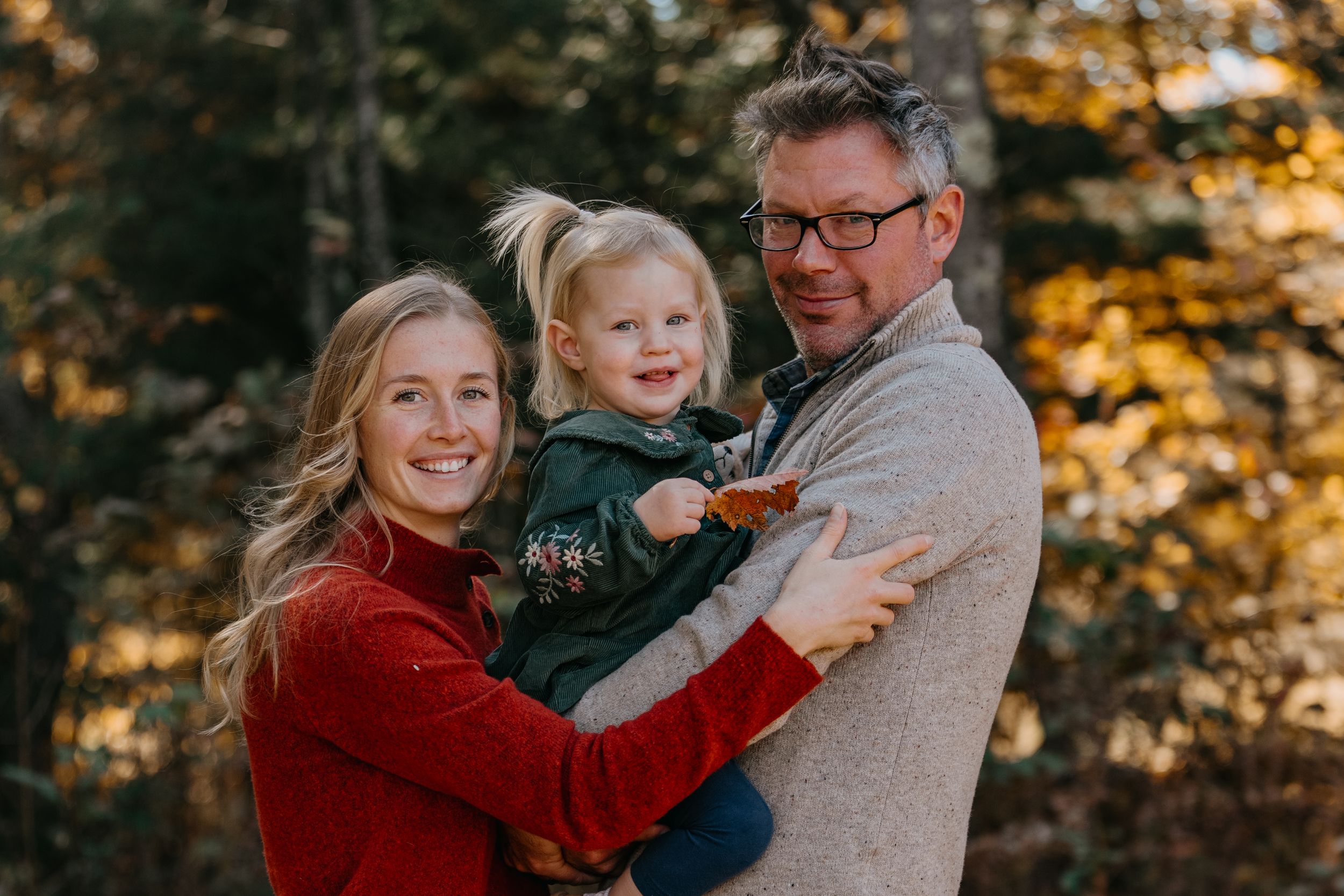 A family shares warm smiles while embracing outdoors during golden hour with autumn foliage in the background.
