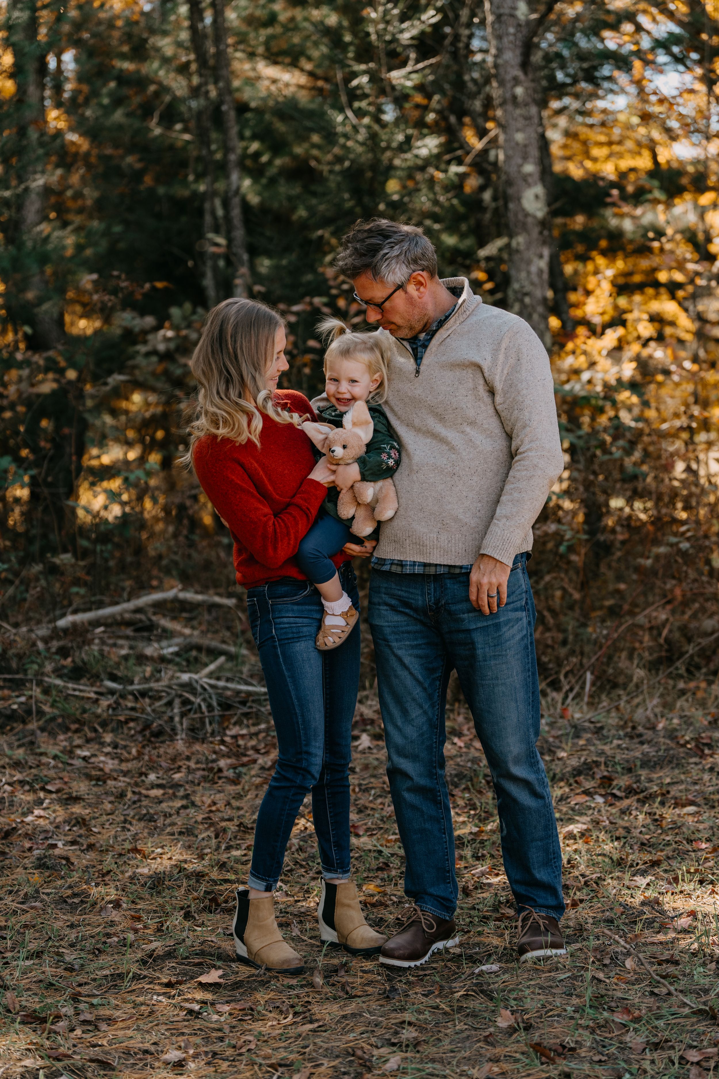 Family in casual fall attire stands together in autumn forest with golden sunlight filtering through trees.
