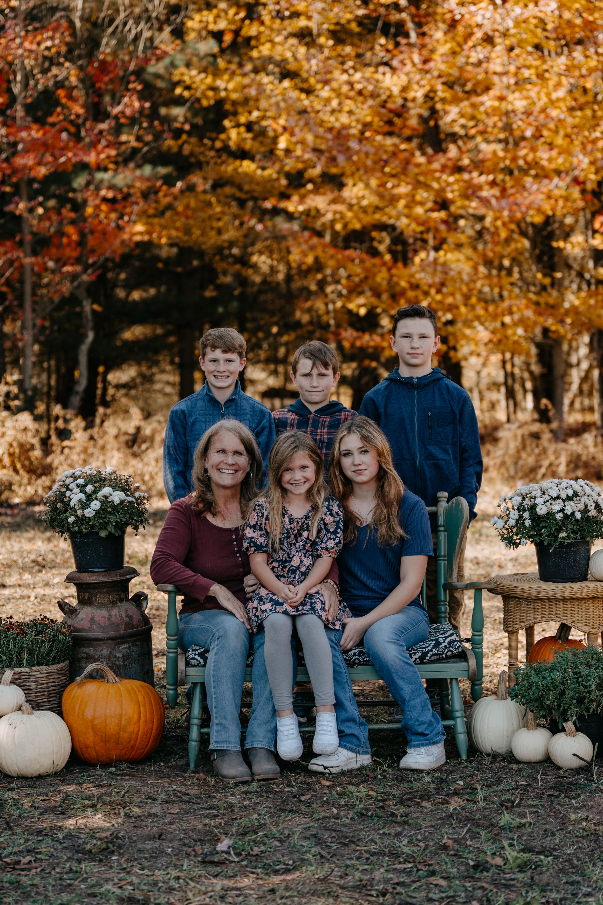Fall family portrait scene with autumn foliage, decorative pumpkins and potted mums surrounding a bench outdoors.