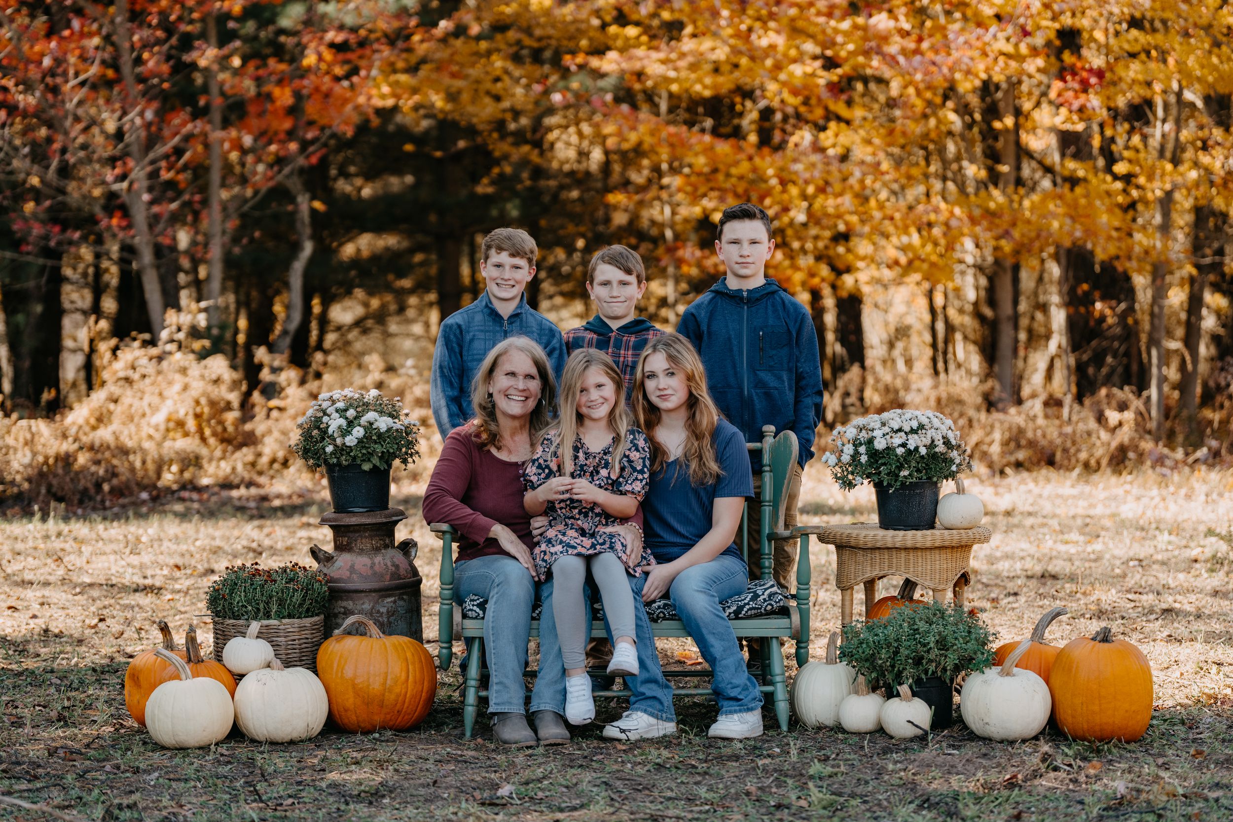 Group sitting outdoors with autumn trees and decorative pumpkins during a fall photo session.